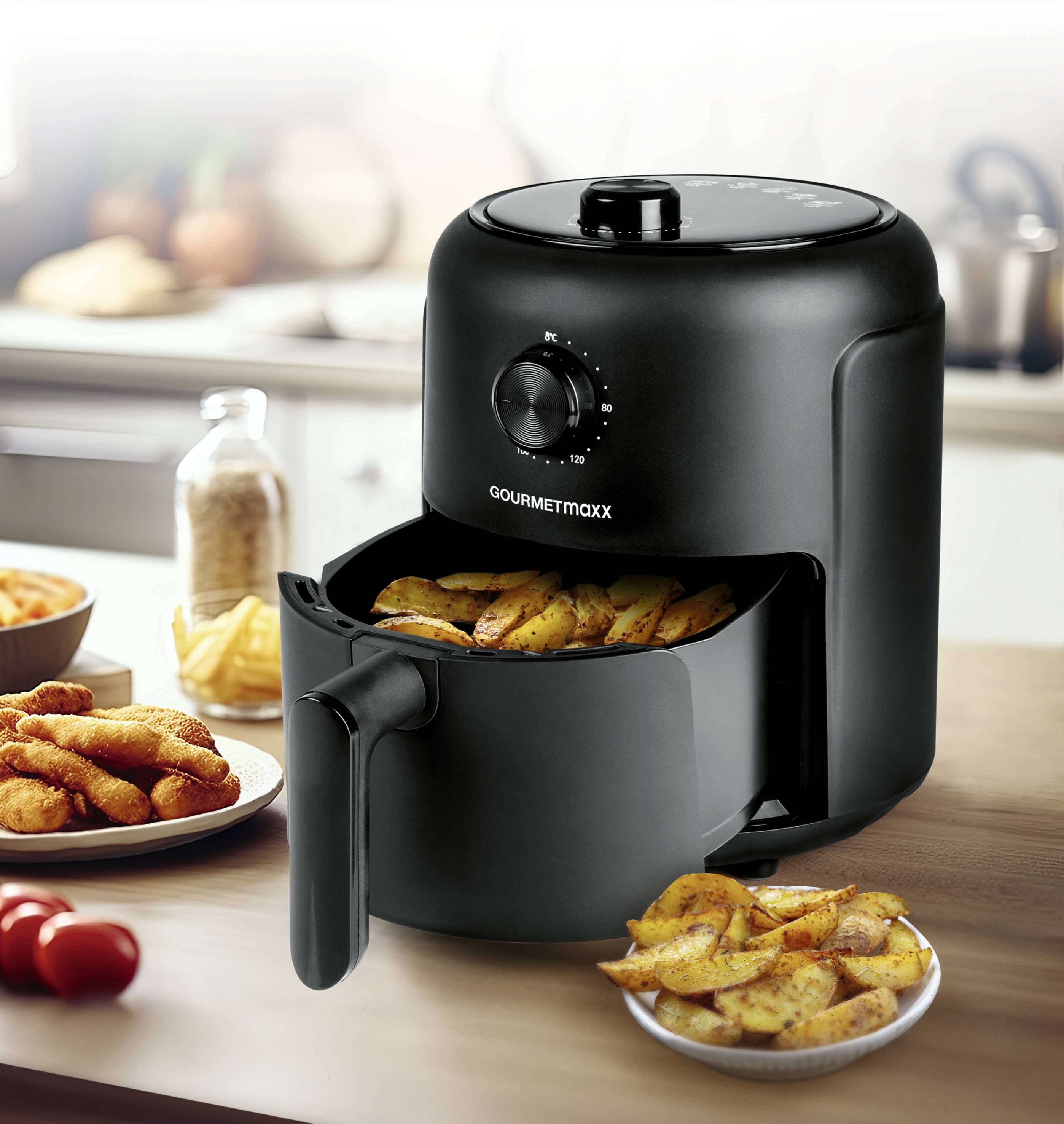 Černá horkovzdušná fritéza na kuchyňském stole, naplněná křupavými hranolkami. V pozadí kuchyňská linka s ingrediencemi.