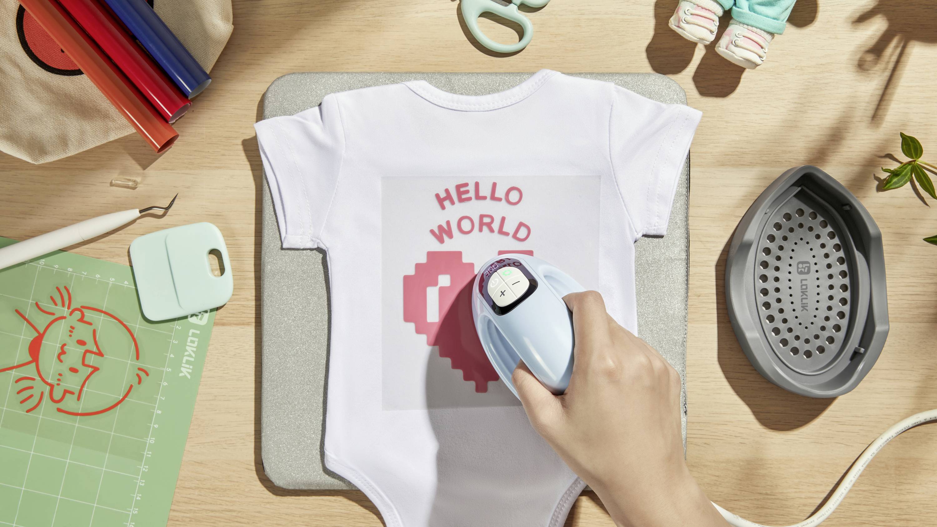 Jedna ruka žehlí dětské body s nápisem 'HELLO WORLD' a motivem srdce. V pozadí jsou viditelné kreativní pomůcky.