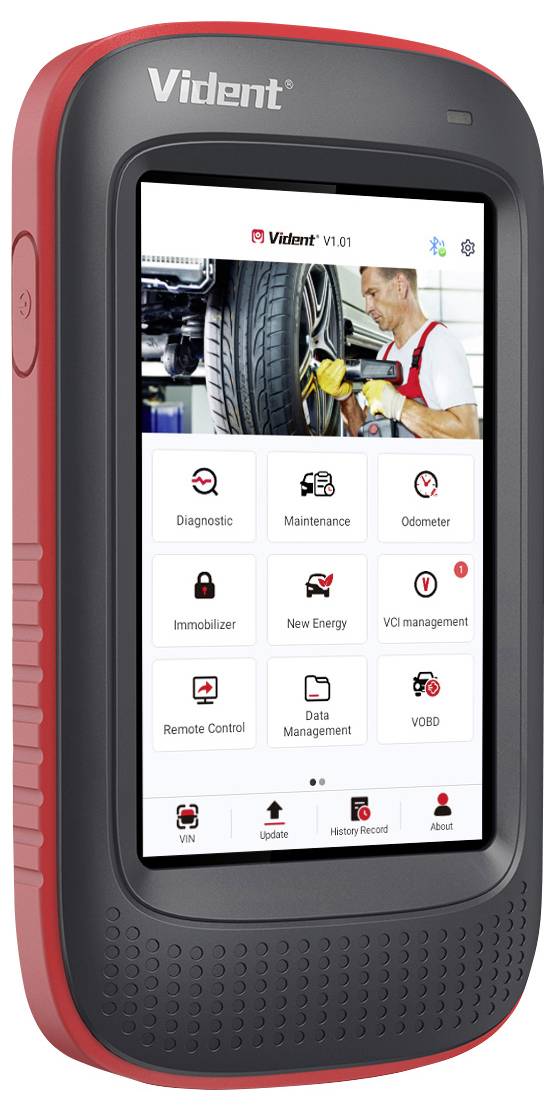 Černý a červený diagnostický nástroj s displejem zobrazuje různé symboly pro diagnostiku a údržbu vozidla. Mechanik pracuje na pneumatice.