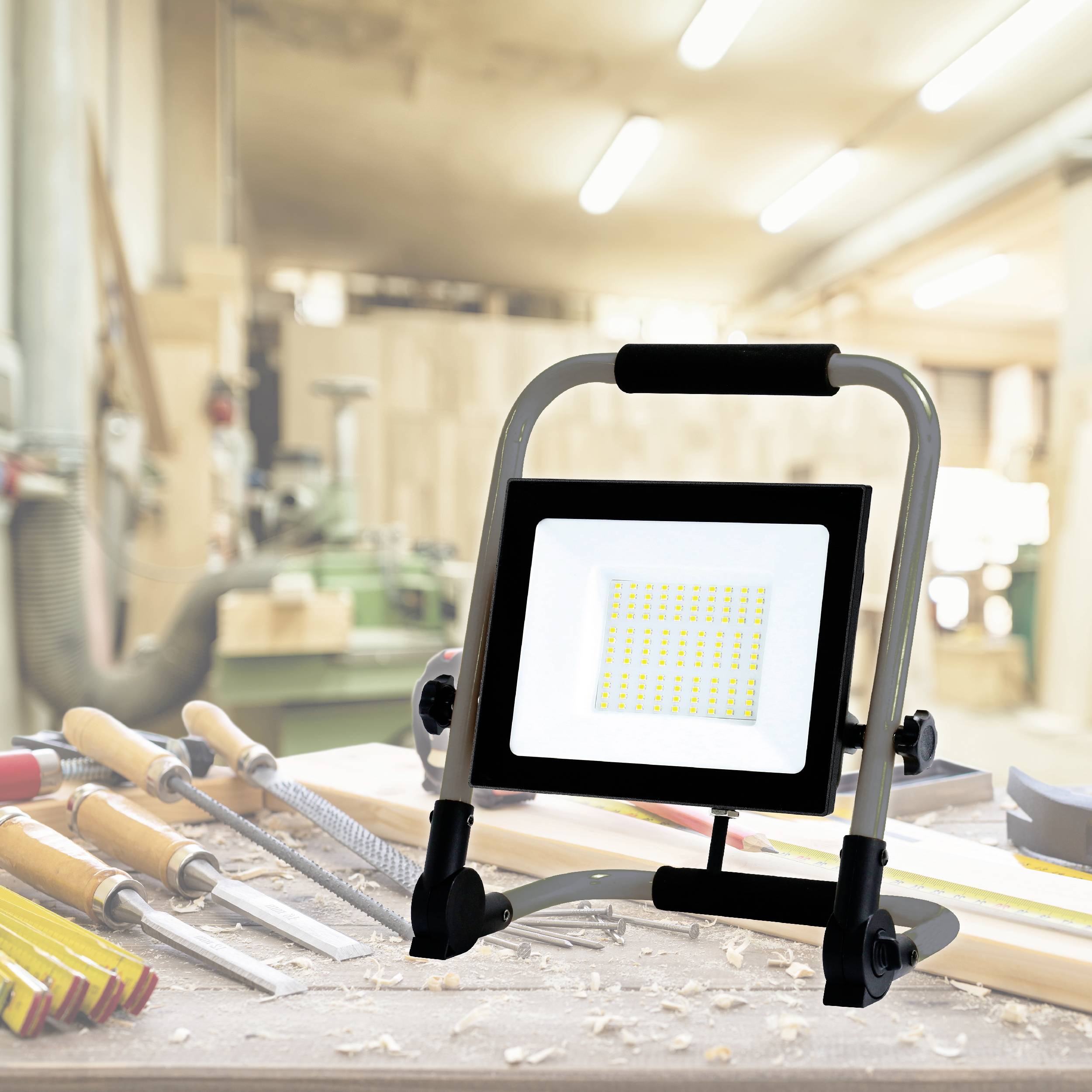 LED reflektor stojí na pracovním stole v dílně. Vedle něj jsou uspořádány různé nástroje jako pilníky a šroubováky.