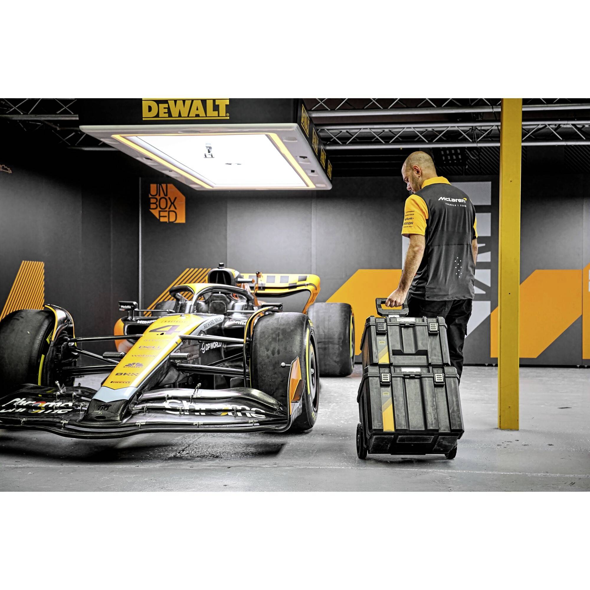 Mechanik táhne vozík s nářadím vedle závodního vozu v dílně. V pozadí je vidět logo společnosti DeWalt.