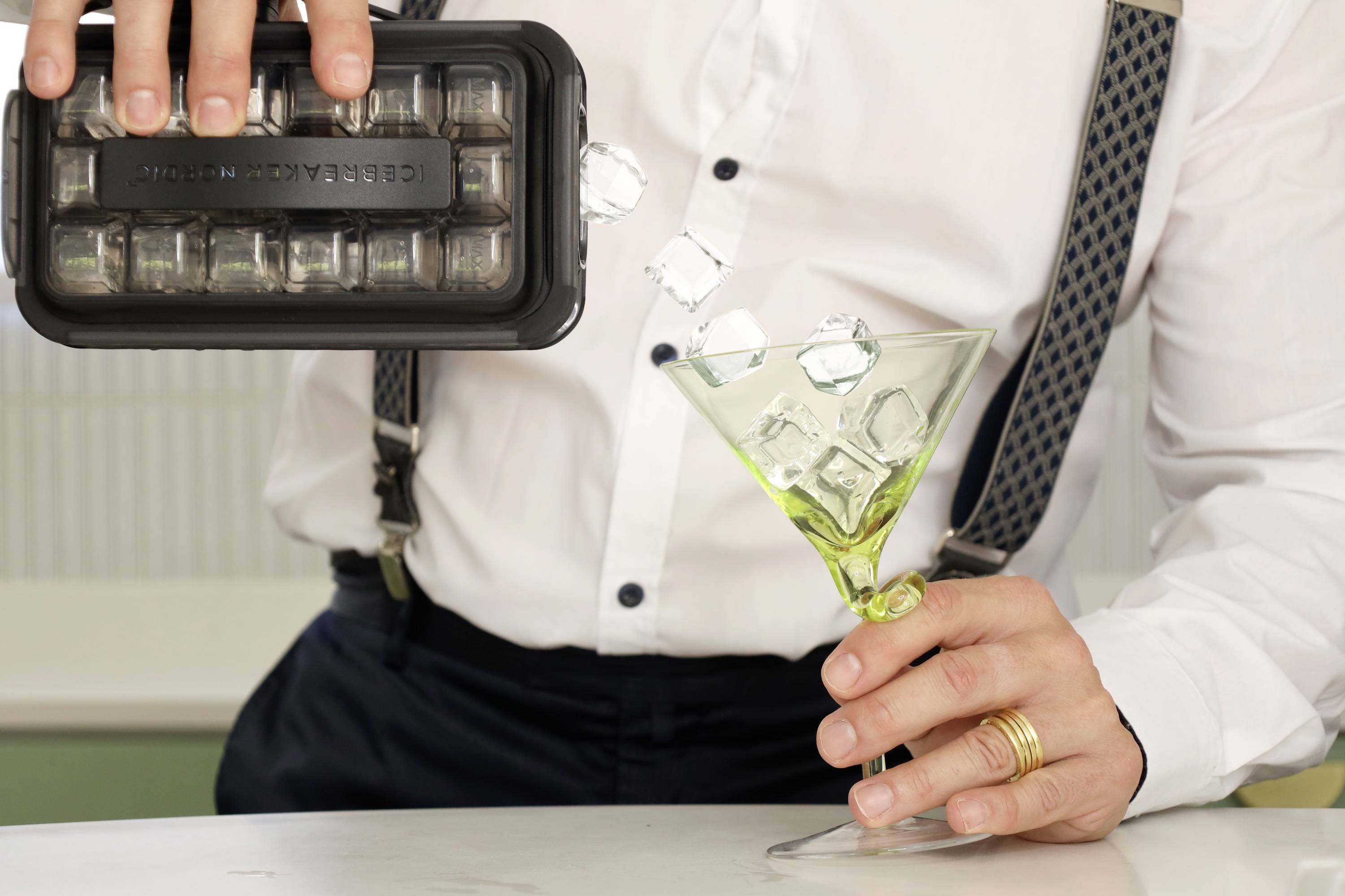 Osoba v bílé košili a na kšandách vyklápí kostky ledu z černé formy do zeleného martini skla.