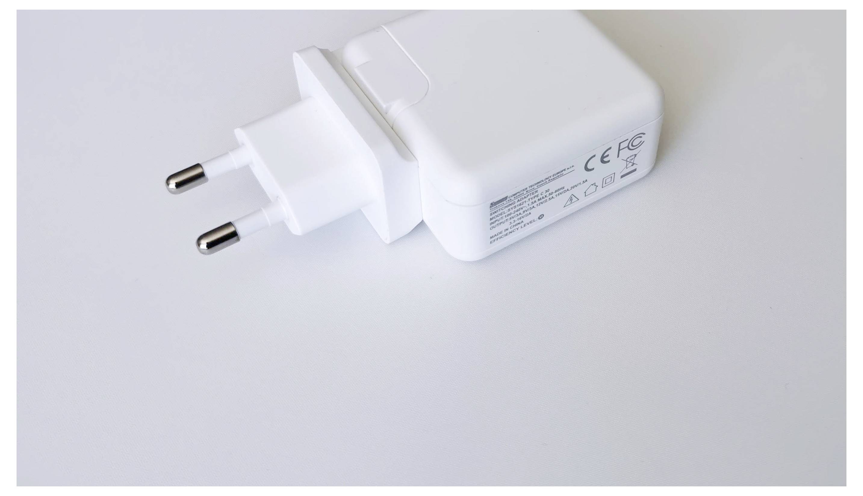 Bílá USB nabíječka s evropskou zástrčkou leží na bílém pozadí. Je viditelný potisk s technickými specifikacemi.