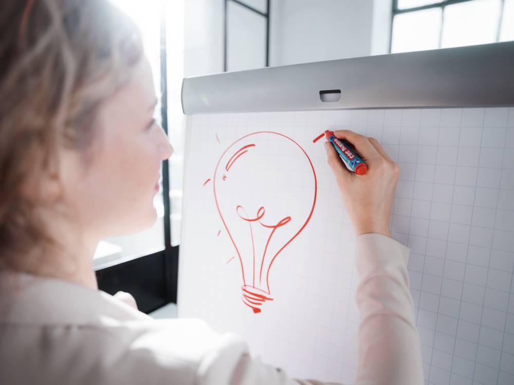 Osoba kreslí červeně zářící žárovku na flipchart. Jasně osvětlená kancelář vytváří kreativní atmosféru.