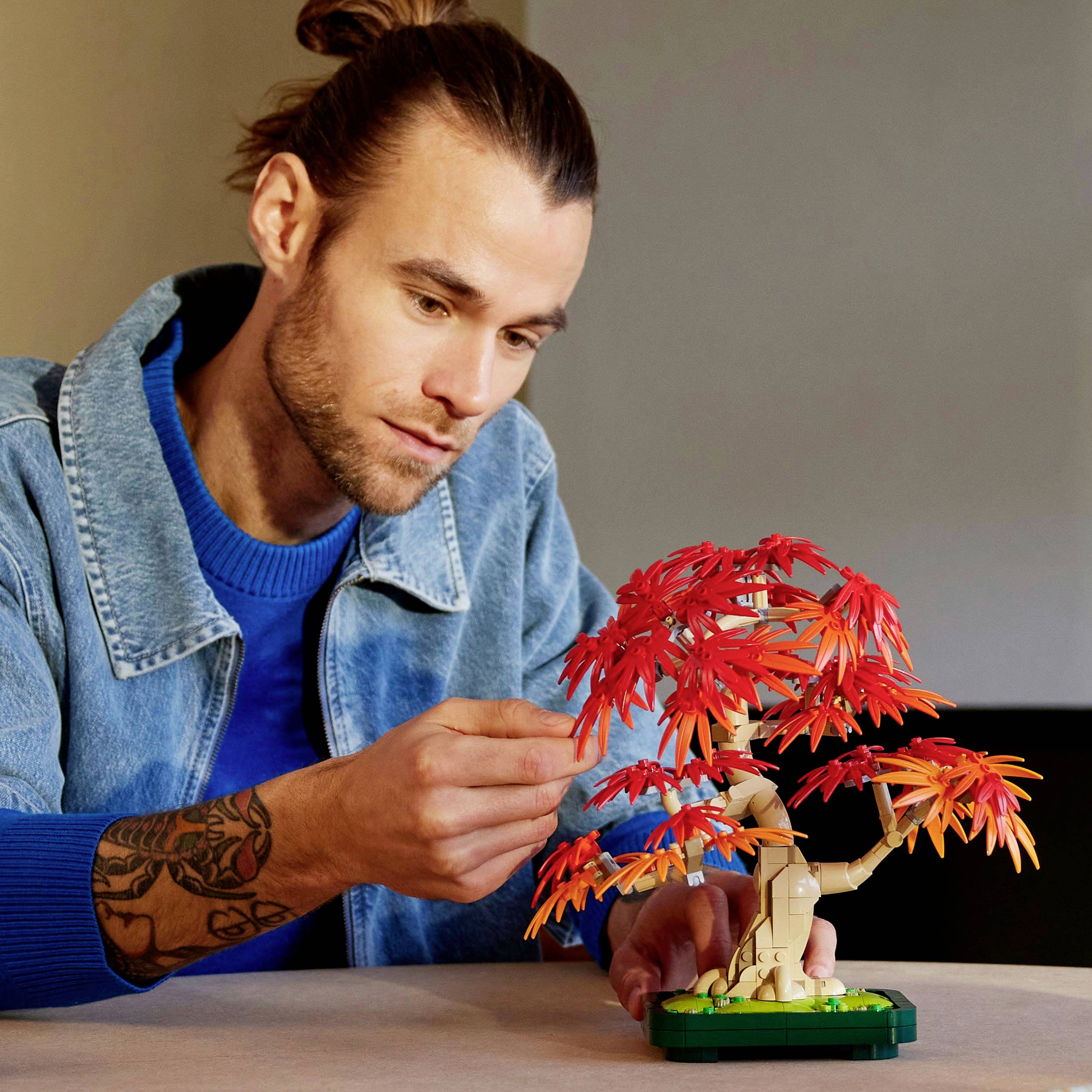 Osoba s tetováními pečuje o malý bonsai strom s červenými listy na stole. Má na sobě džínovou bundu a modrý svetr.