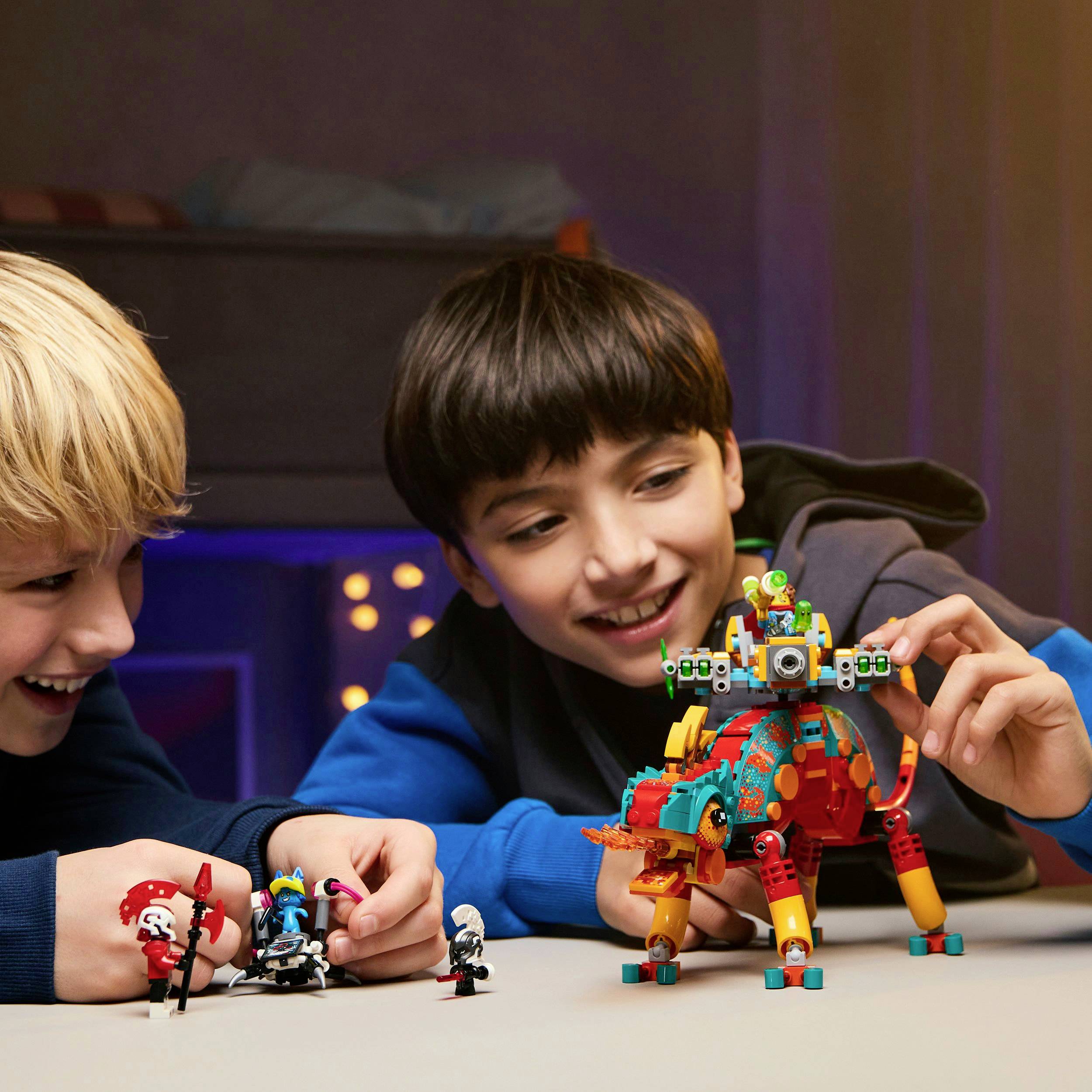 Dvě děti si hrají s barevnými modely robotů na stole. Usmívají se a soustředí se na své konstrukce.