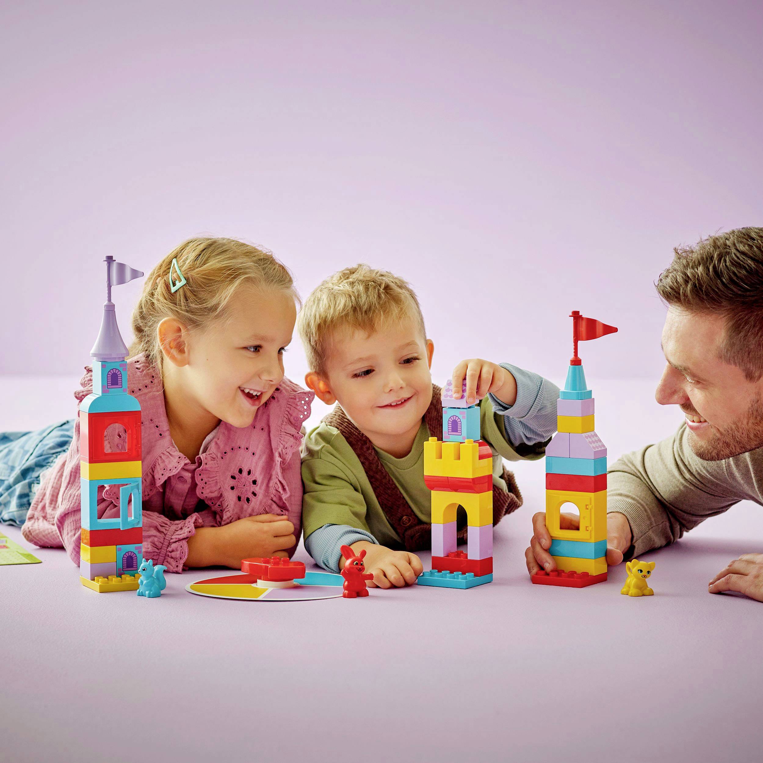 Děti si hrají s barevnými stavebními kostkami, staví věže a společně se smějí. Dospělý je pozoruje a zapojuje se do hry.
