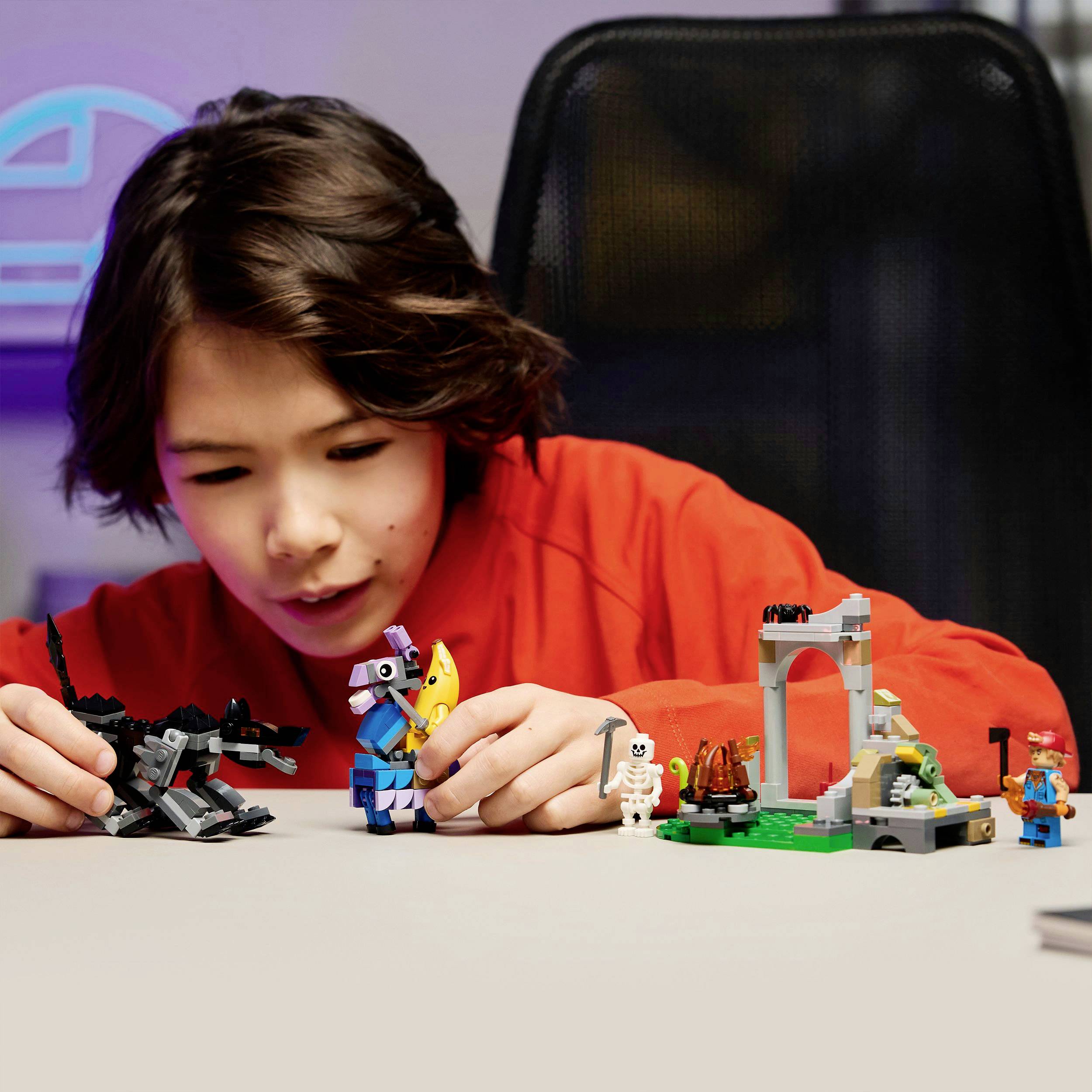 Dítě si hraje s kostkami Lego na stole. Drží figurku a sedí obklopené různými konstrukcemi z Lega.