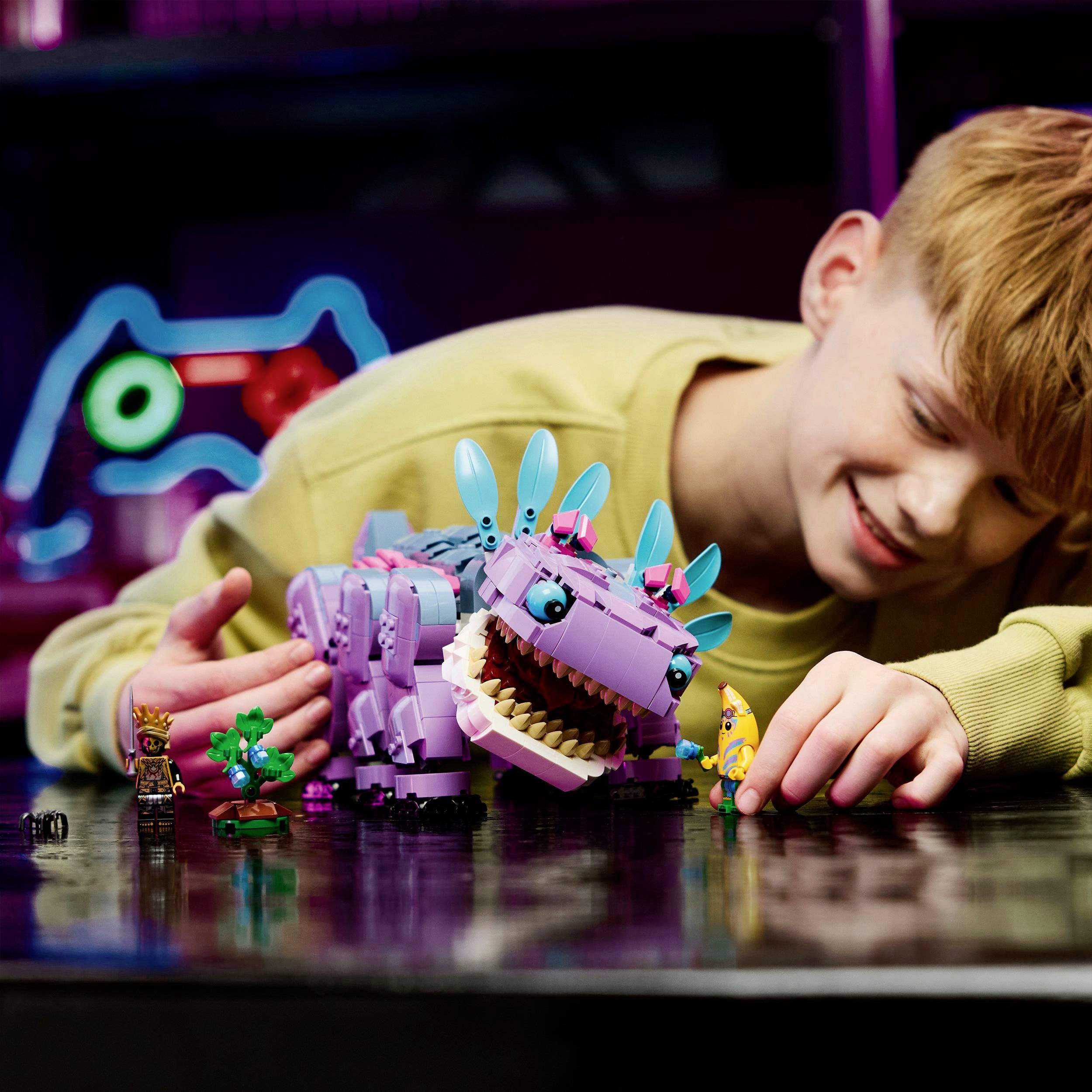 Dítě si s úsměvem hraje s fialovým dinosauřím hračkou, obklopené barevnými figurkami na stole.