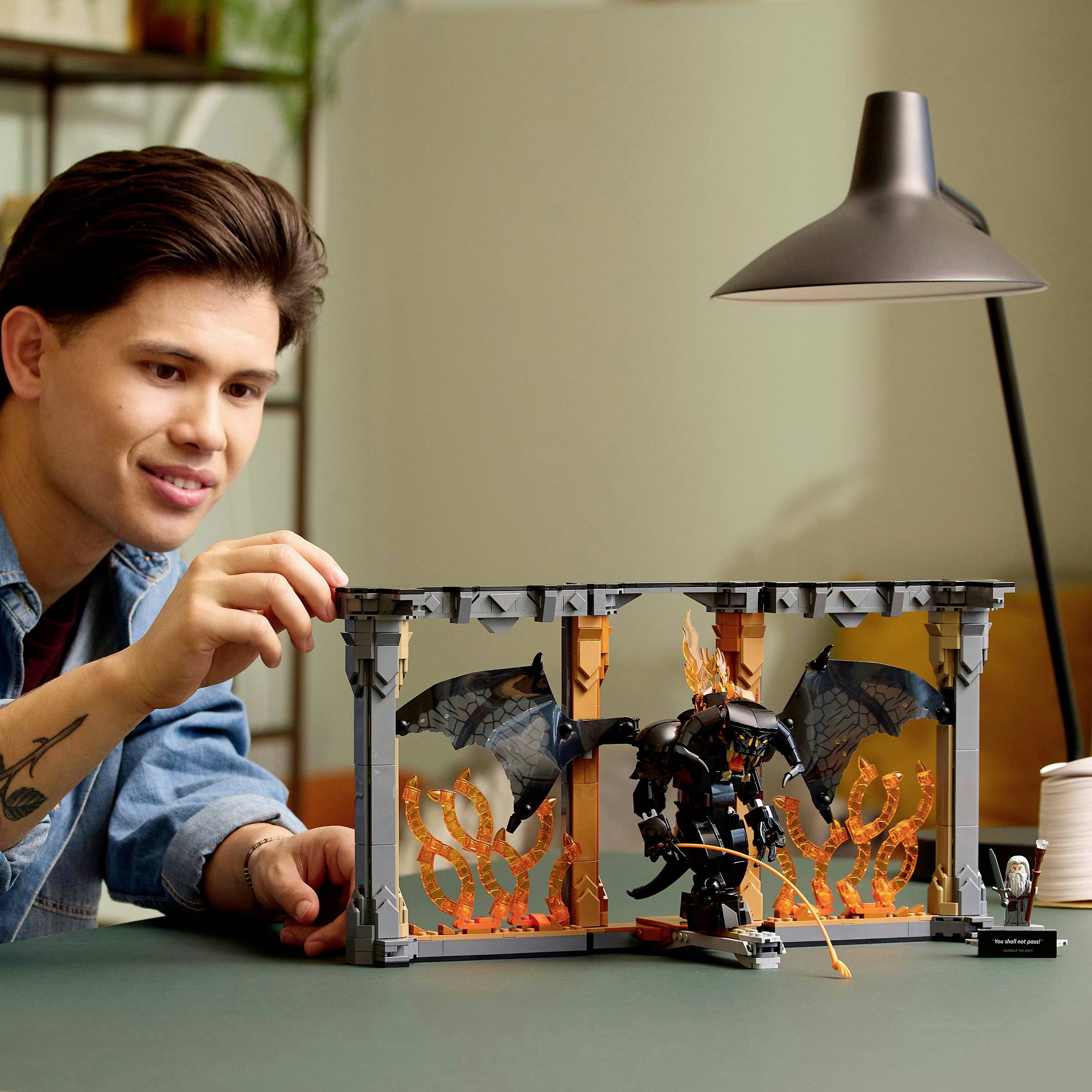 Mladý muž soustředěně pozoruje Lego stavebnici s figurkou, která má černá křídla a oranžové plameny, na stole.