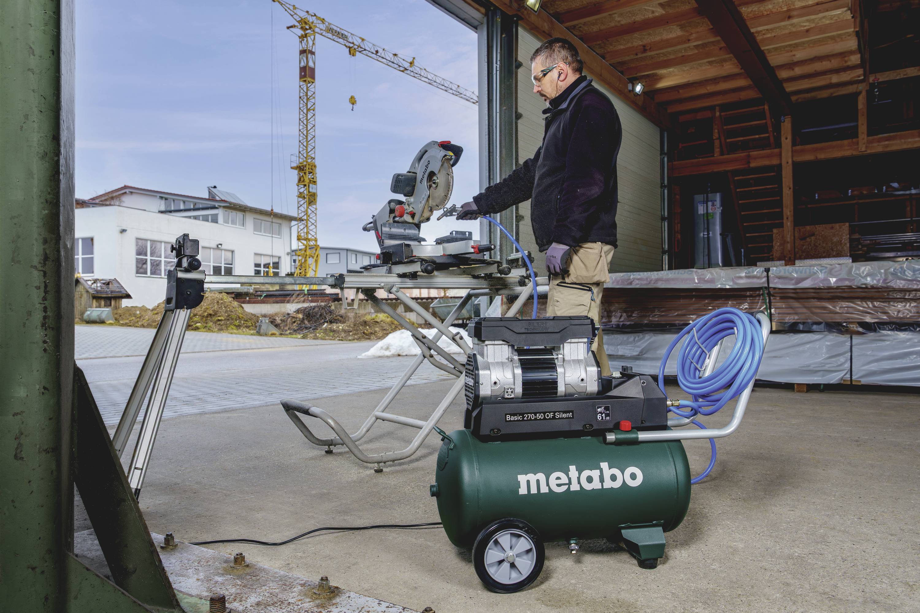 Osoba obsluhuje elektrickou pilu v dílně. V popředí stojí zelený kompresor značky 'Metabo'.