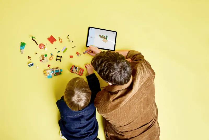 Dítě a dospělý sestavují hračku na žluté ploše podle pokynů zobrazených na tabletu.