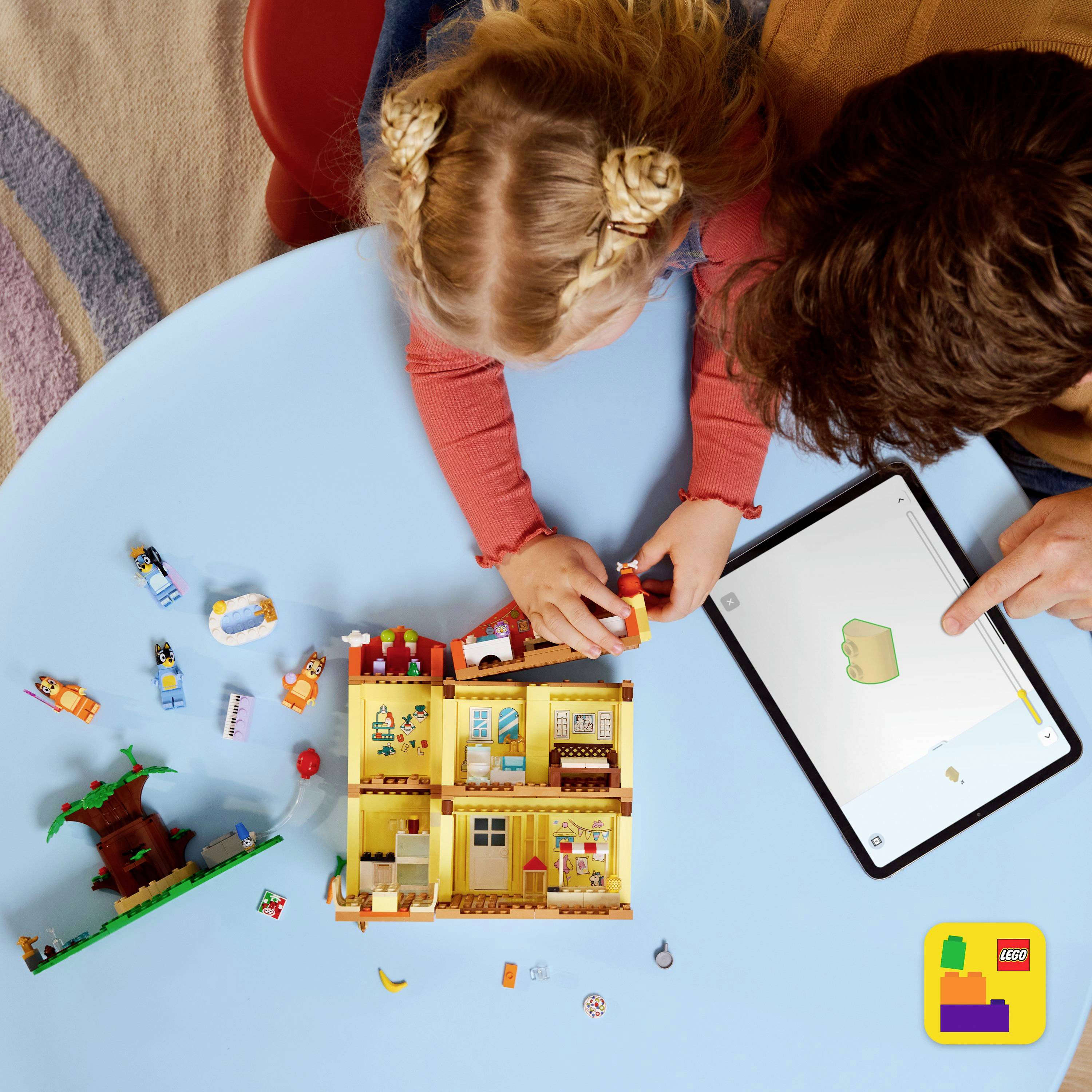 Dítě si hraje s LEGO, zatímco dospělý ukazuje související návrh na tabletu. Sada LEGO připomíná malý dům s nábytkem a figurkami.