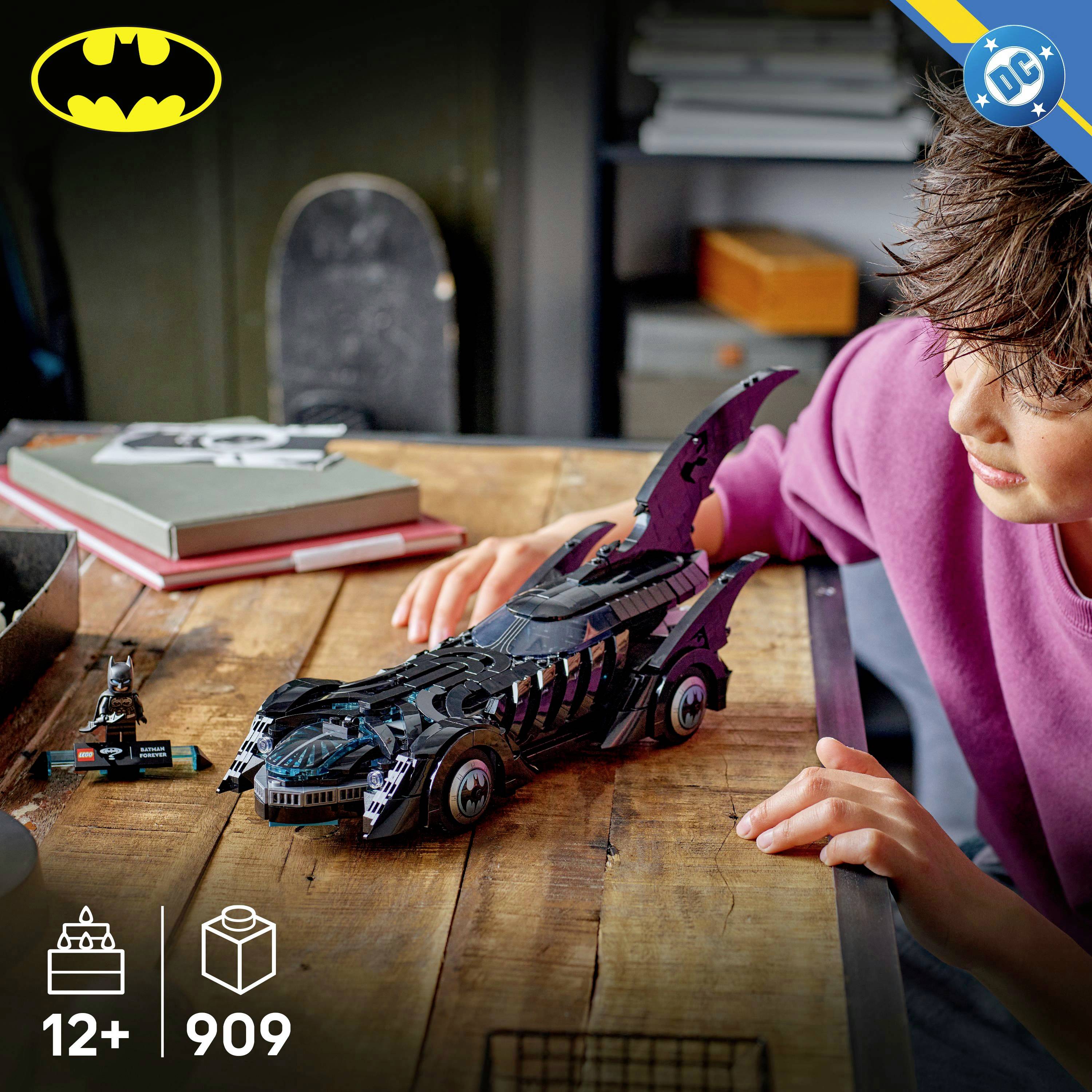 Dítě v růžovém tričku si hraje s detailně zpracovaným Lego Batmobilem na dřevěném stole, poblíž je malá figurka Batmana.