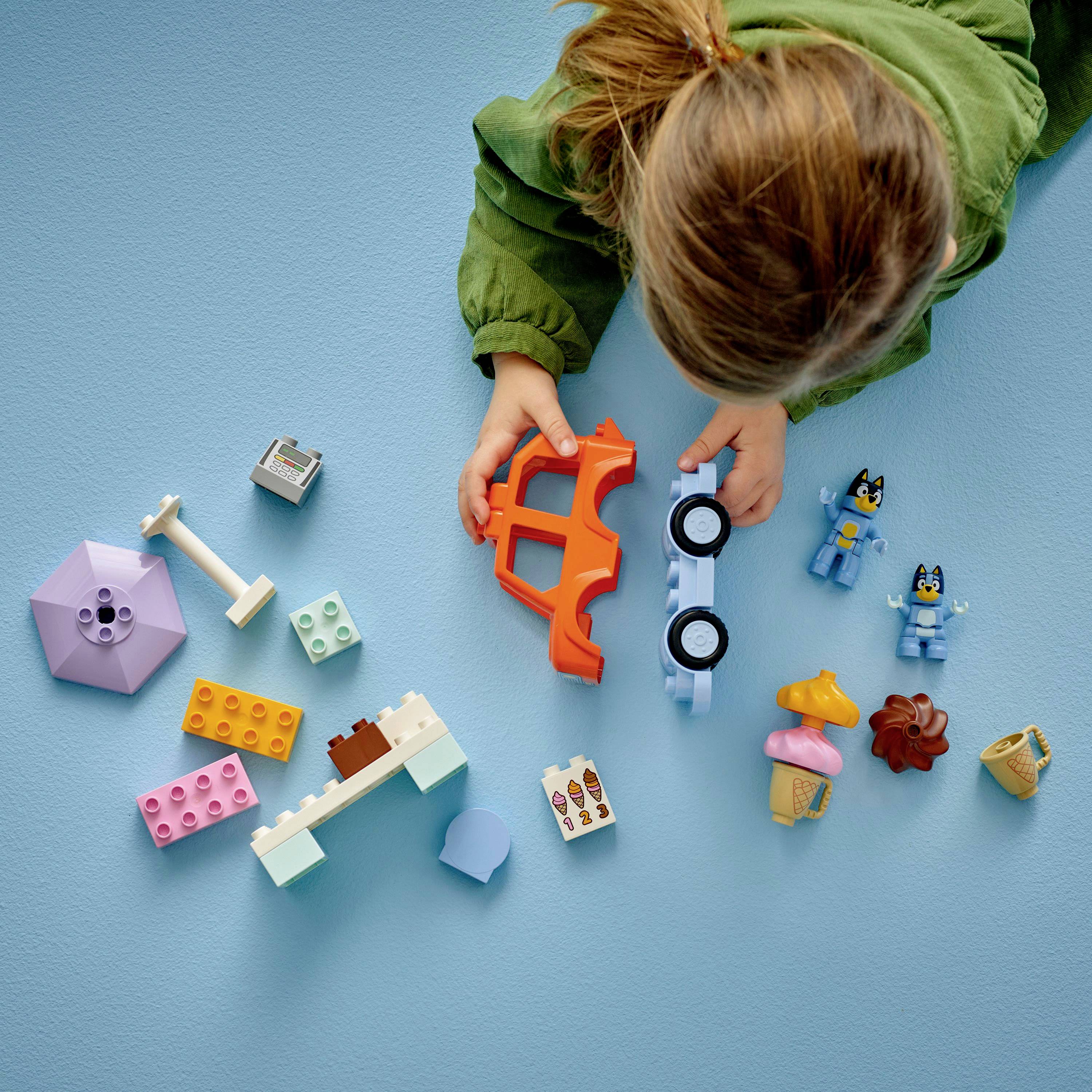 Dítě v zelené bundě si hraje na modré podlaze s různobarevnými hračkami, včetně stavebních kostek, figurek, autíčka a malých zvířátek.