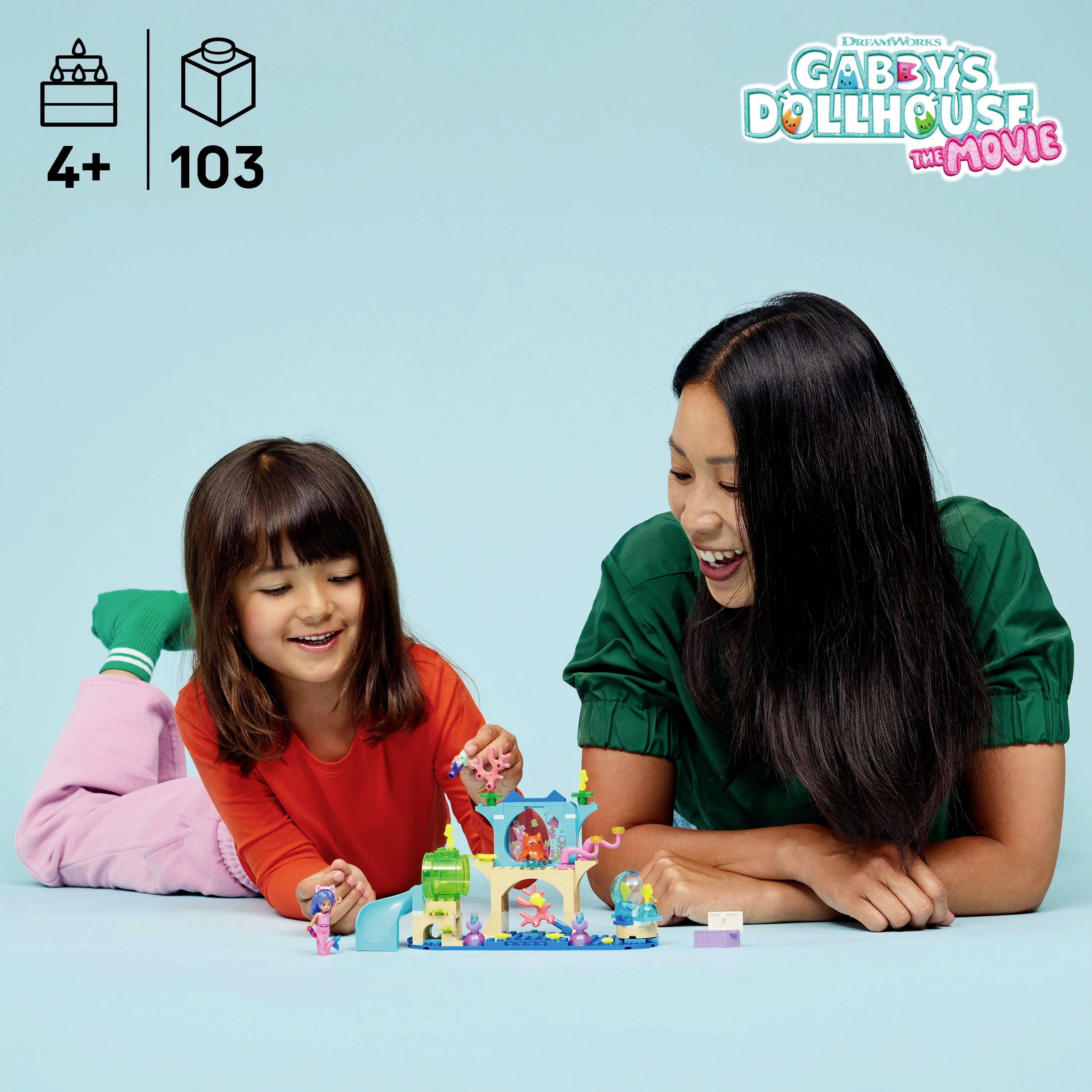 Žena a dítě společně šťastně staví LEGO soupravu 'Gabby's Dollhouse' na podlaze, s barevnými hračkami a zámkem.