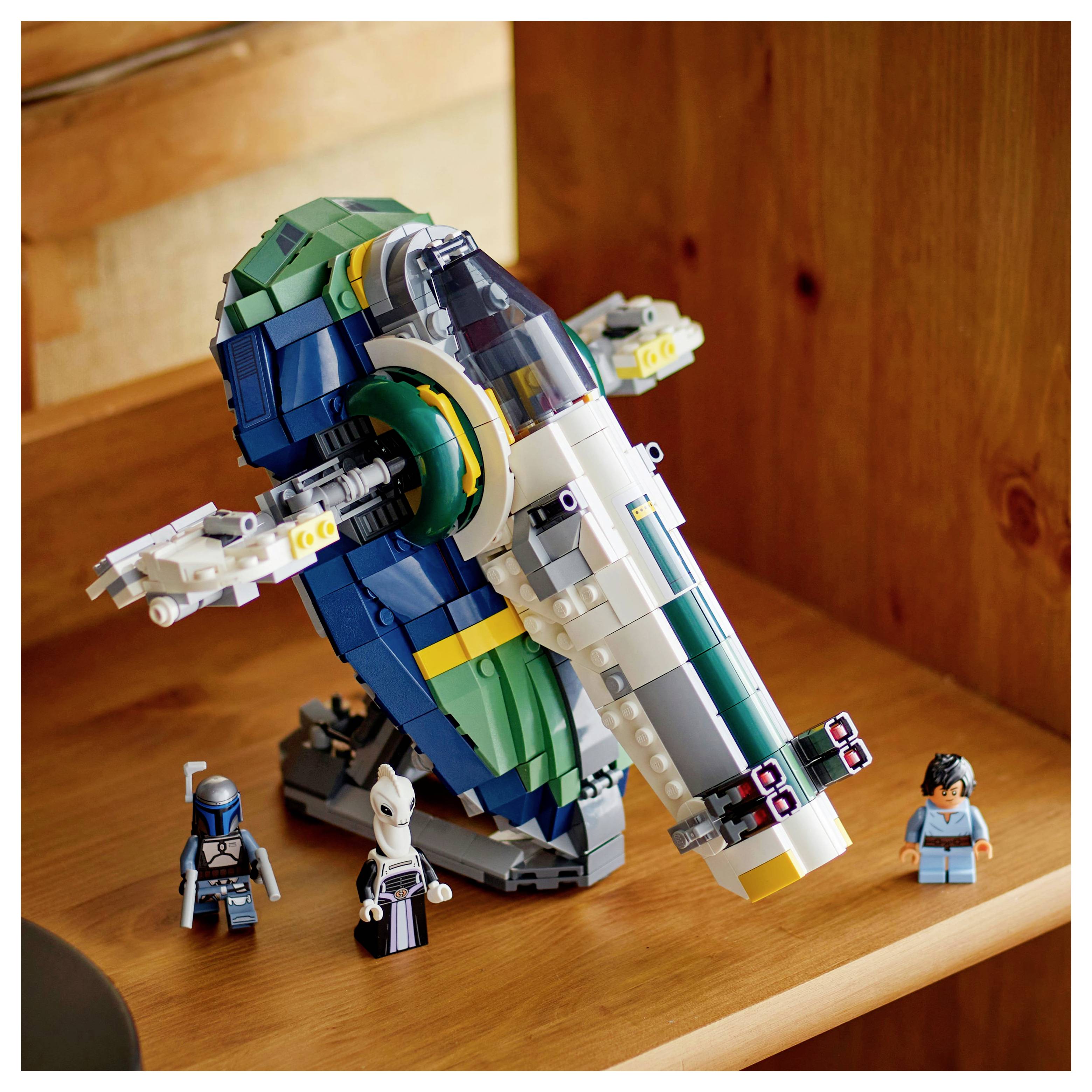 Barevný model vesmírné lodi z kostek LEGO se třemi LEGO figurkami vystavený na dřevěné polici, který se vyznačuje detailním, zakřiveným designem.