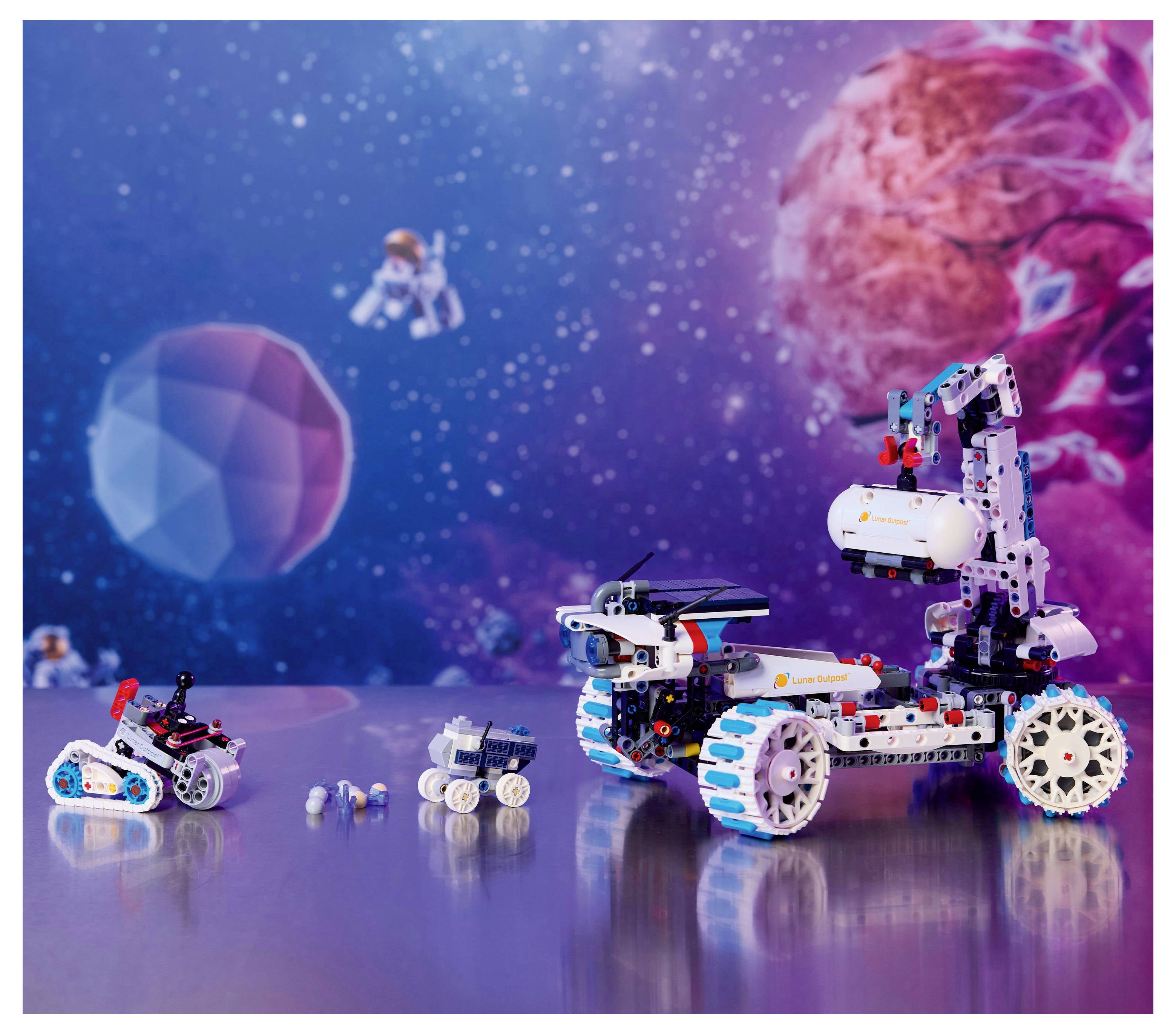 Robotické modely jsou vystaveny na kovovém povrchu s barevným vesmírným pozadím zobrazujícím planety a astronauty.