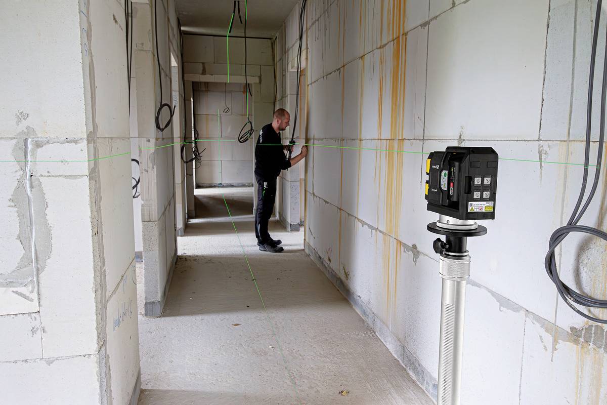 Stavební dělník používá laserový nivelační přístroj k měření stěn rozestavěné budovy. Kabely visí ze stropu.