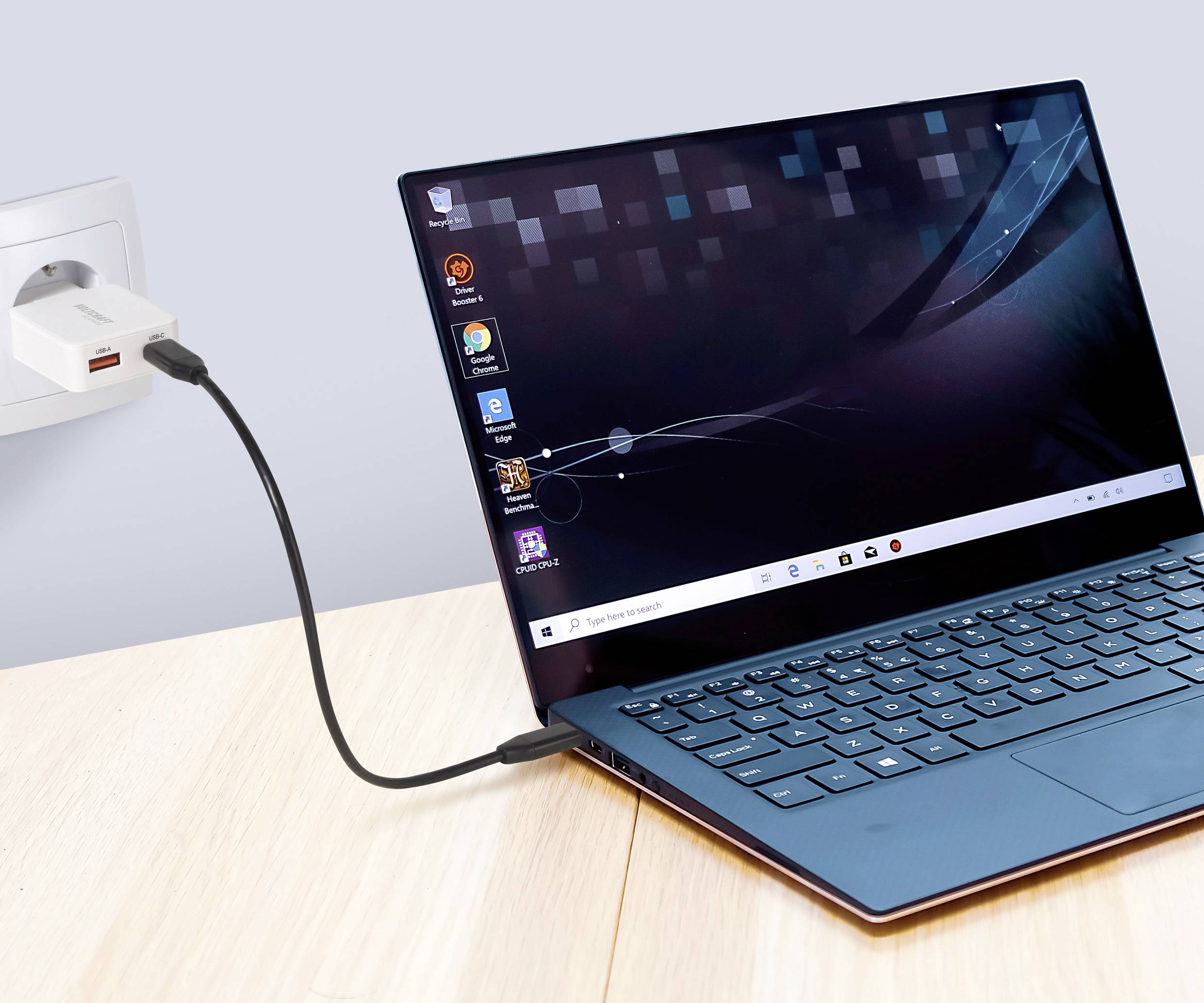 Modrý laptop nabíjející se na dřevěném stole, připojený do elektrické zásuvky ve zdi. Na obrazovce jsou zobrazené různé aplikace na ploše s tapetou na pozadí.