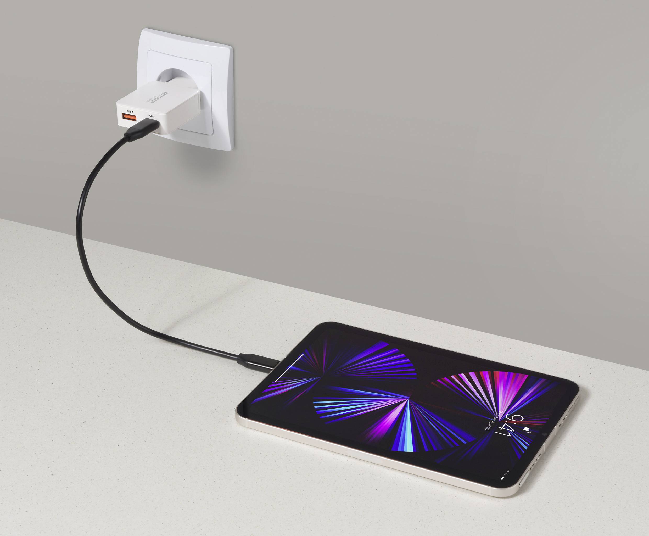 Tablet nabíjející se na pracovní desce, připojený k elektrické zásuvce pomocí nabíjecího kabelu a adaptéru USB-C. Tablet zobrazuje živý abstraktní design.