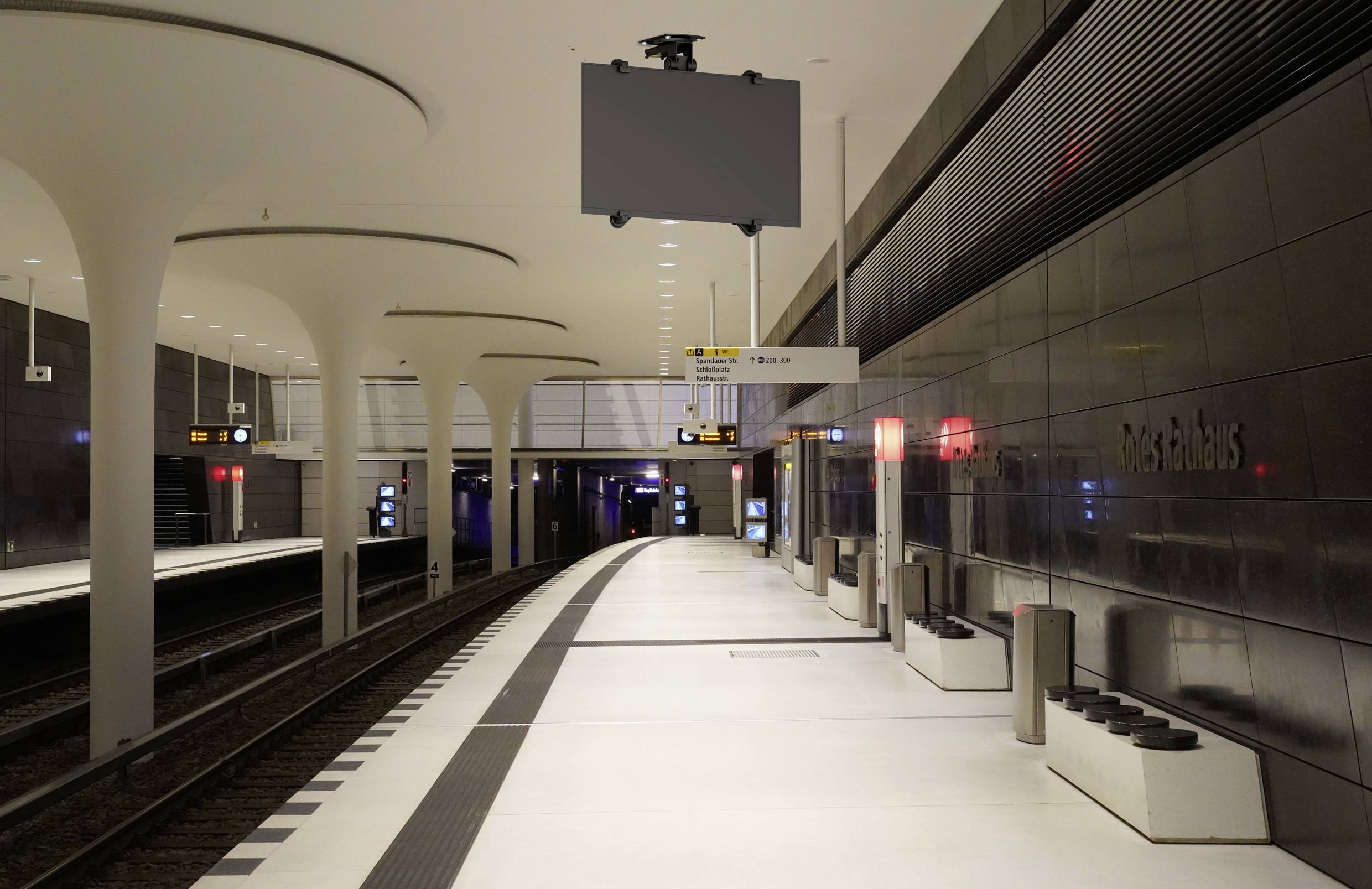 Prázdná moderní stanice metra s bílou podlahou, vysokými sloupy a tmavými stěnami. Koleje jsou na levé straně, nikde není vidět žádná osoba.