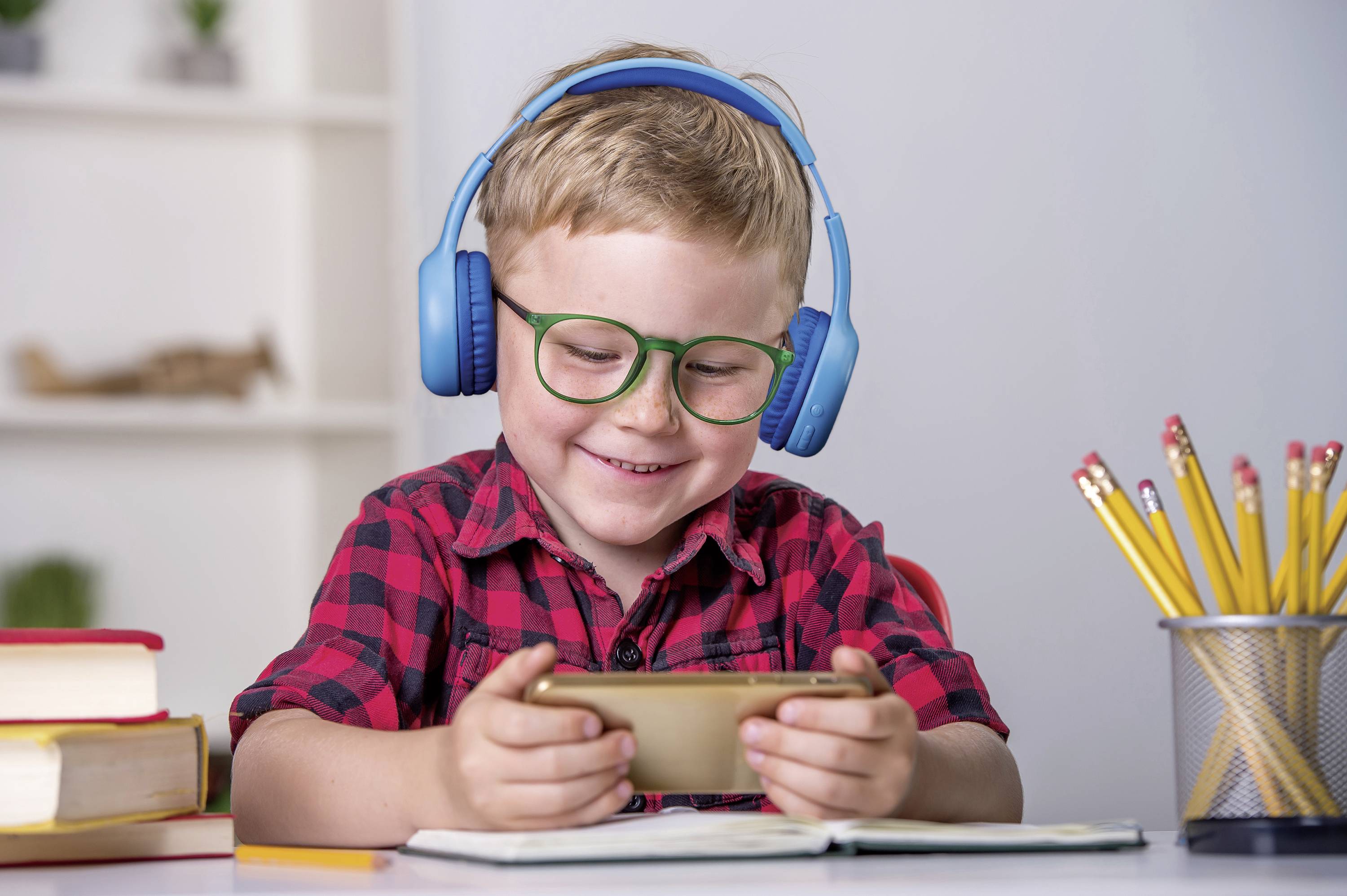 Usměvavý chlapec s brýlemi a modrými sluchátky hraje na smartphone u stolu. Vedle něj leží knihy a stojánek na tužky.