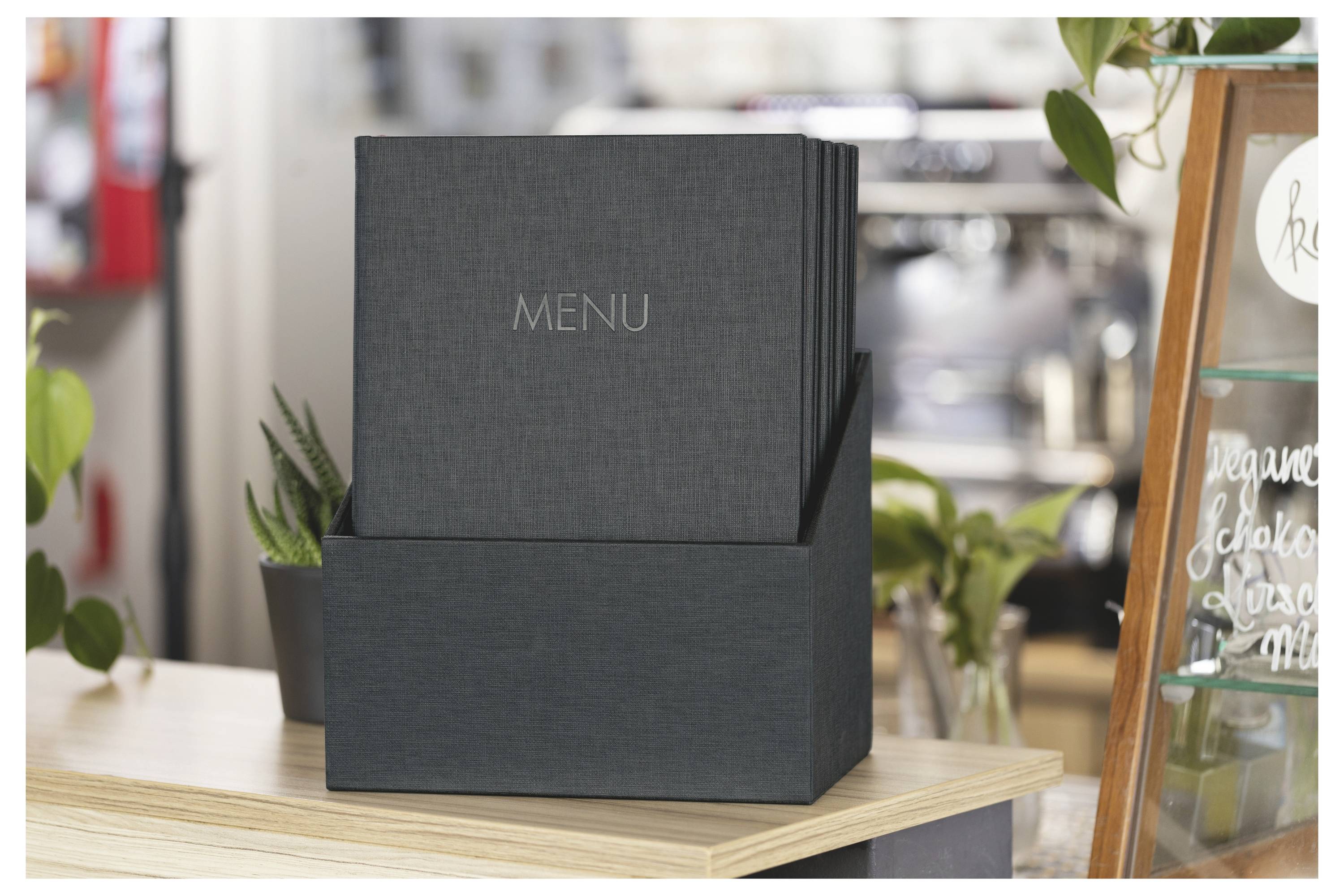 Černá krabice s nabídkou na prodejním pultu kavárny, označená "MENU", v pozadí s rostlinami a tabulí napsanou křídou.