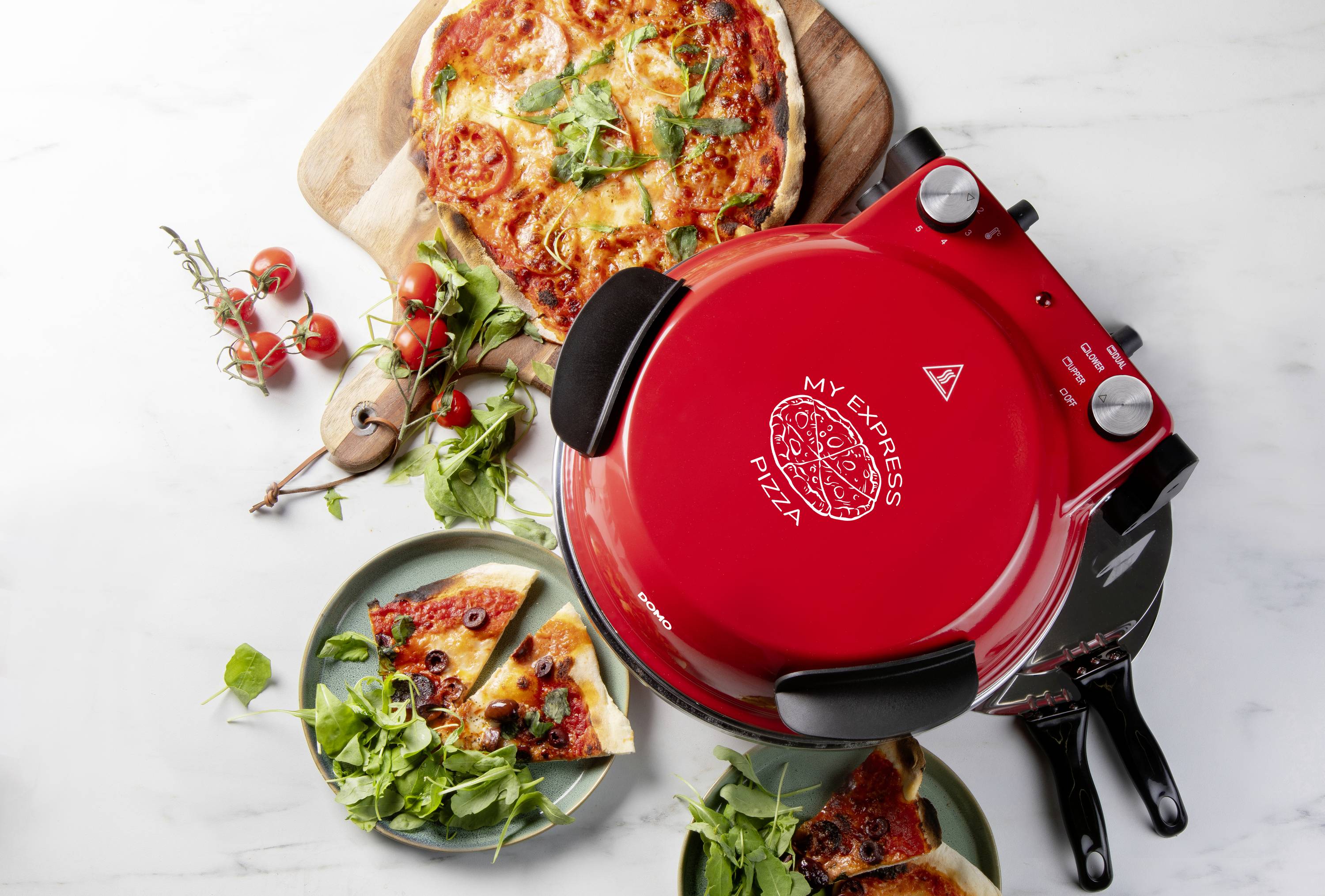 Červená pizová pec s hotovou upečenou pizzou vedle ní. Čerstvé rajčata a bylinky leží na prkénku vedle pece.