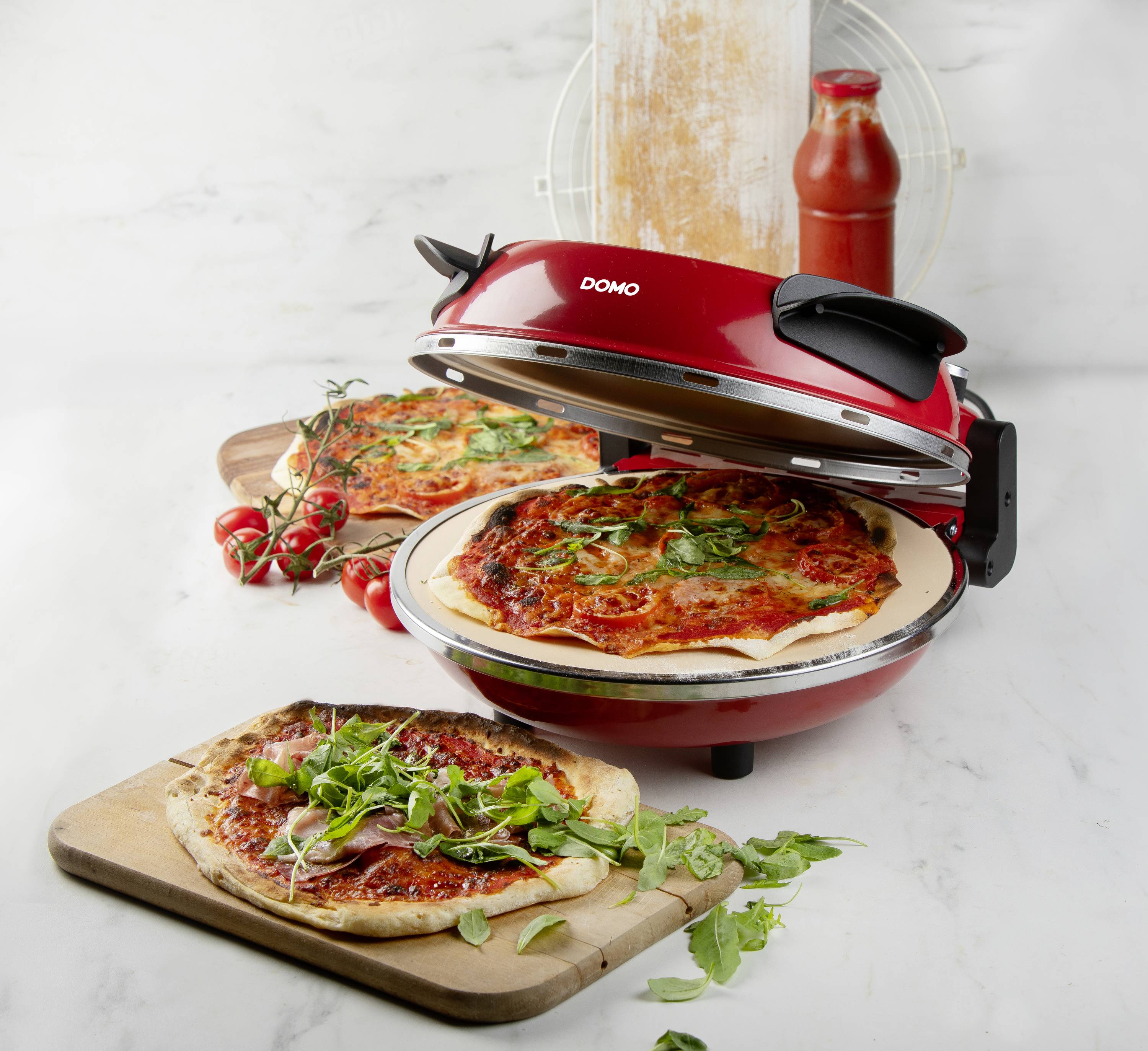 Červená pizza pec vedle dvou čerstvých pizz na dřevěné desce. Pizzy jsou ozdobené rajčaty, bylinkami a sýrem. V pozadí láhev s omáčkou.