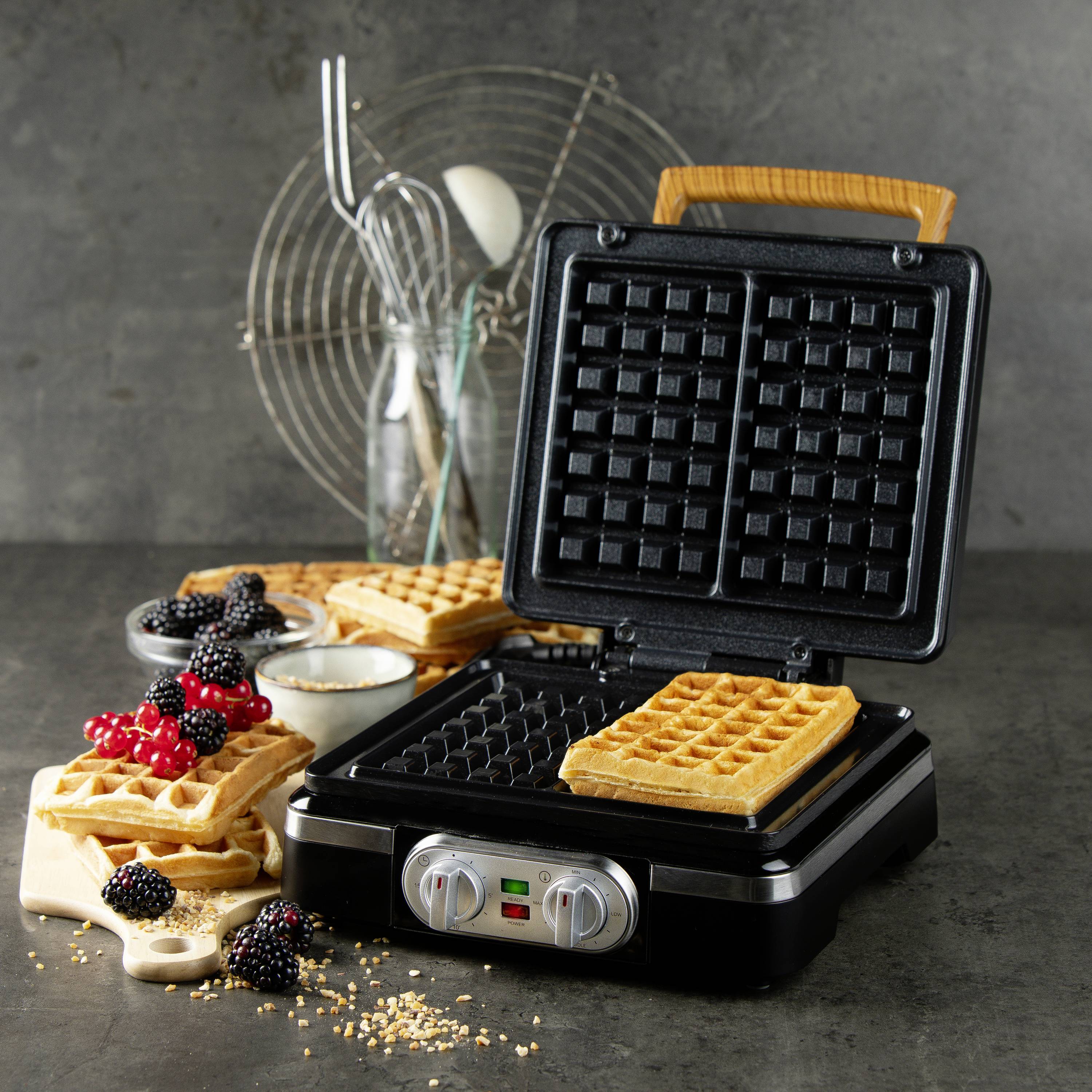 Waflová forma s čerstvě upečenou wafkou vedle ní. Bobulové ovoce, šlehačka a další wafle jsou uspořádány na prkénku.