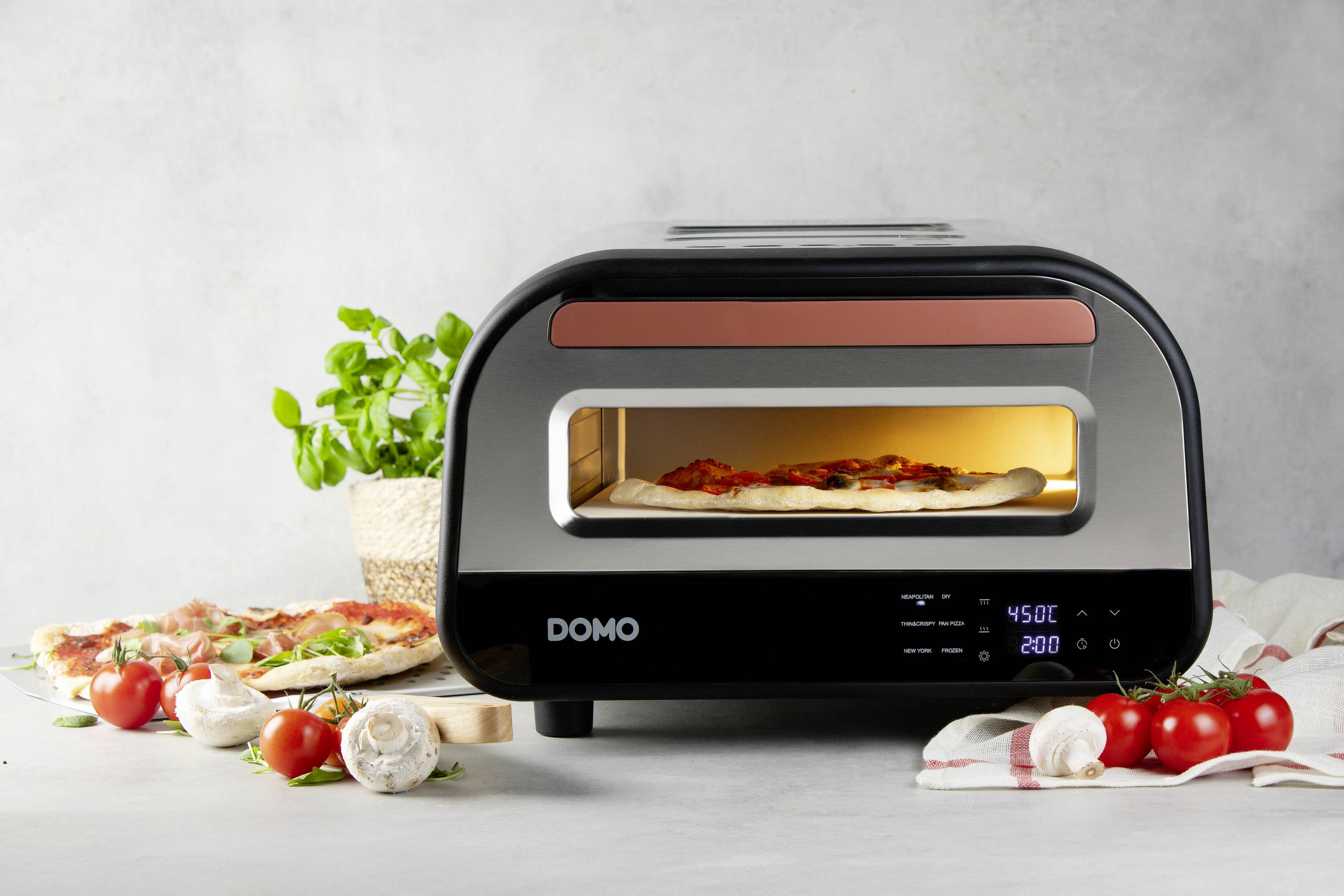 Moderní pizza pec s pizzou uvnitř, obklopená čerstvými ingrediencemi jako jsou rajčata a bylinky na kuchyňské pracovní desce.