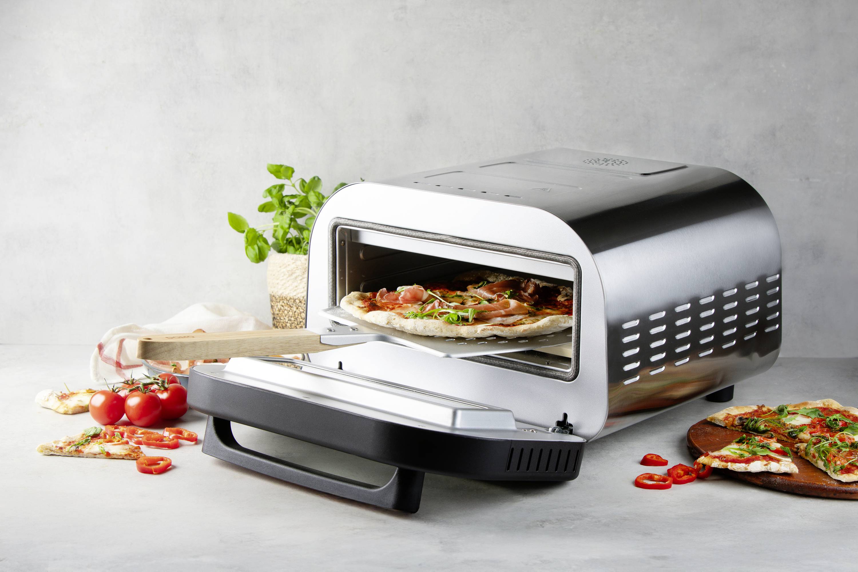 Stříbrná pec na pizzu s otevřenými dvířky a dřevěnou lopatou, na které leží pizza. Vedle jsou čerstvé rajčata, bylinky a nakrájená pizza.