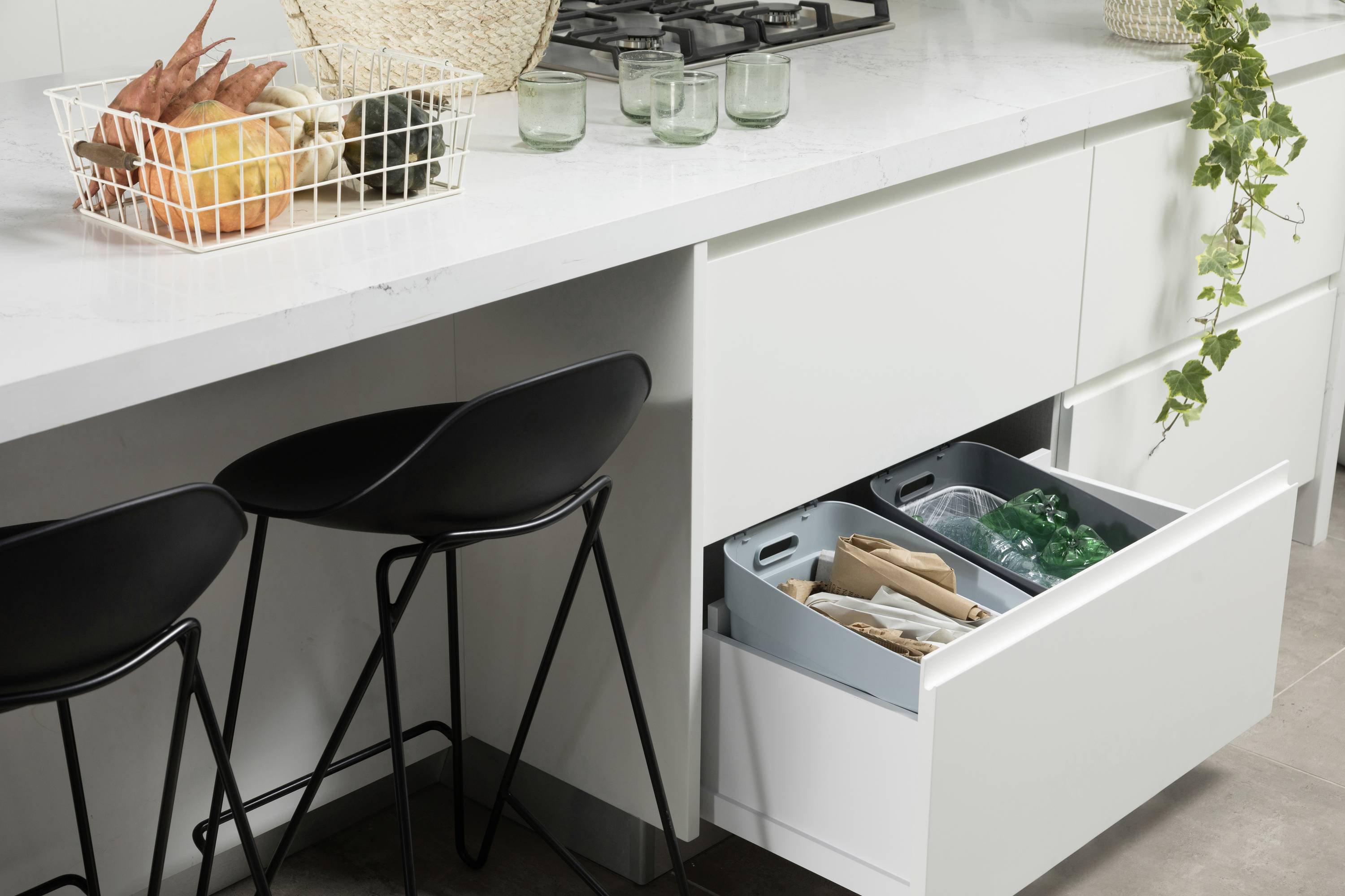 Dva černé barové stoličky stojí před bílým kuchyňským ostrovem. Jedna zásuvka je otevřená a uvnitř jsou vidět recyklované materiály.
