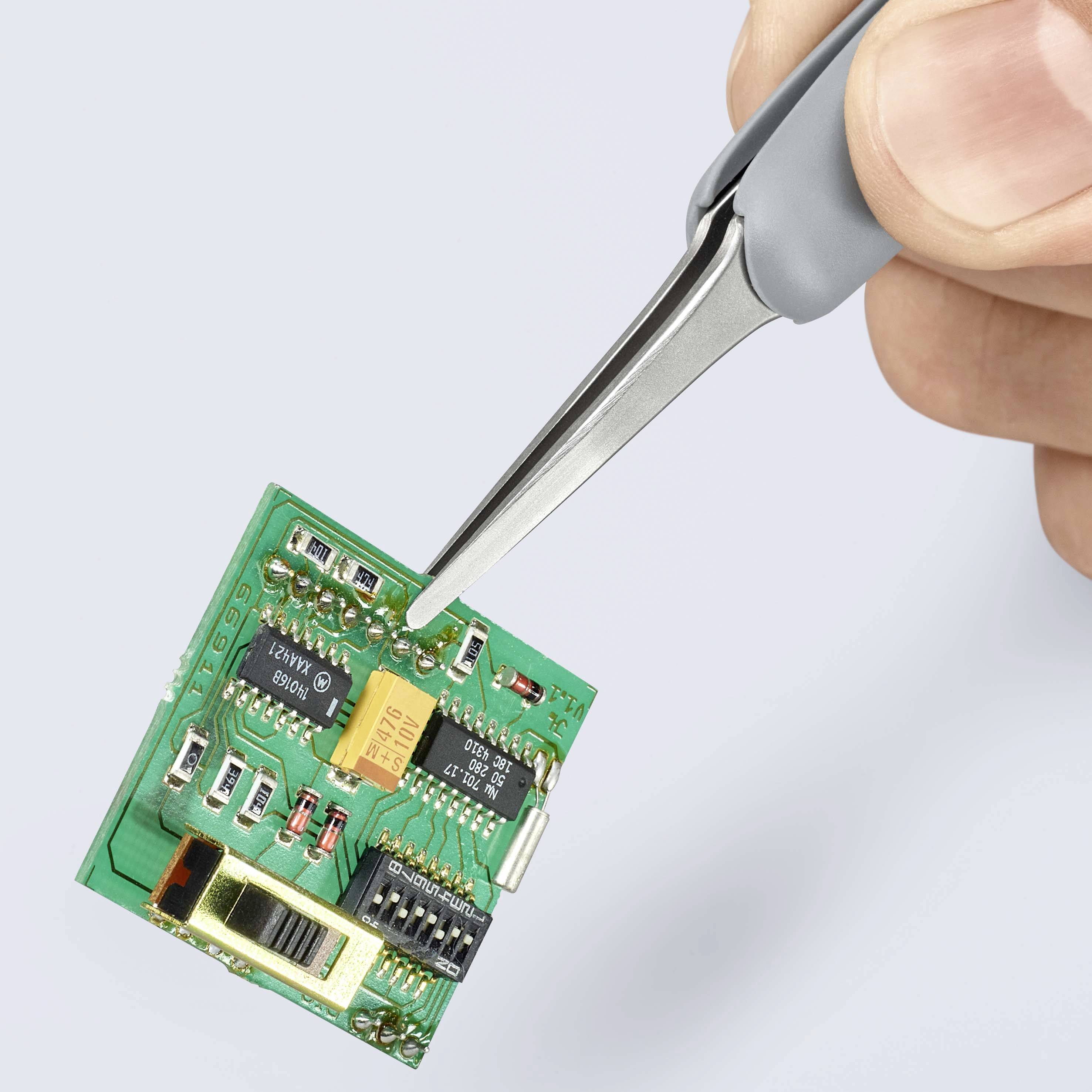 Jedna ruka drží pinzetou zelenou elektronickou desku plošných spojů osázenou různými komponenty.
