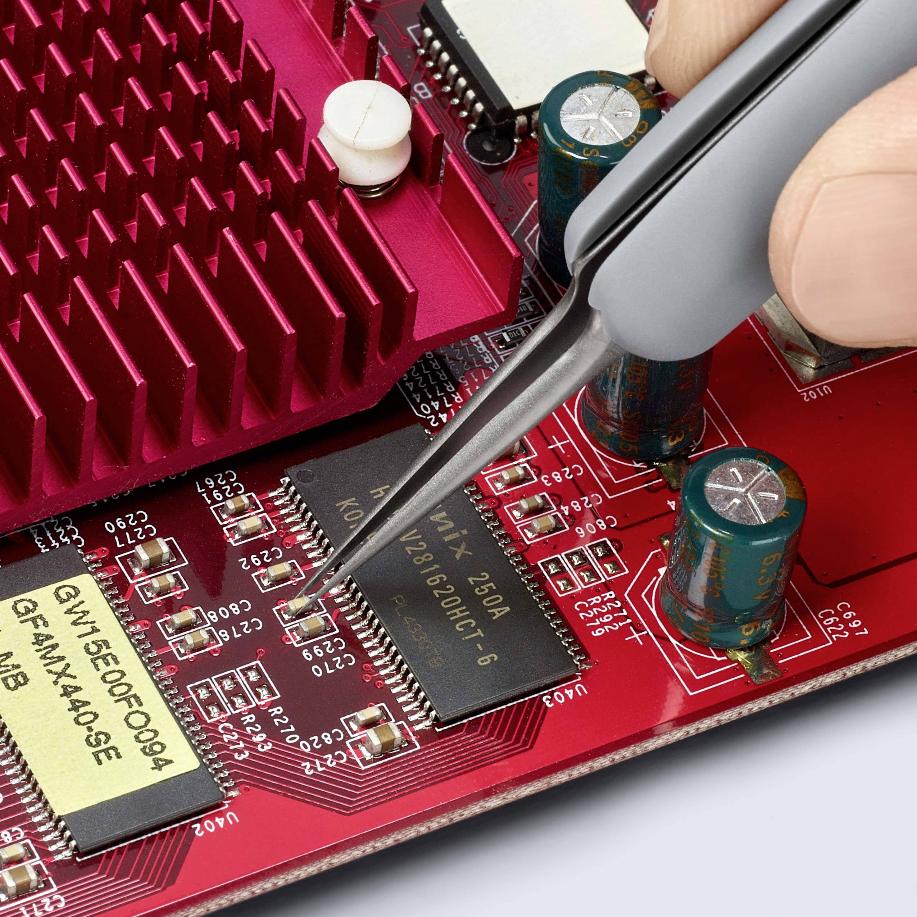 Jedna ruka drží pinzetu a umisťuje mikrochip na červenou desku plošných spojů s viditelnými obvody a elektronickými komponenty.