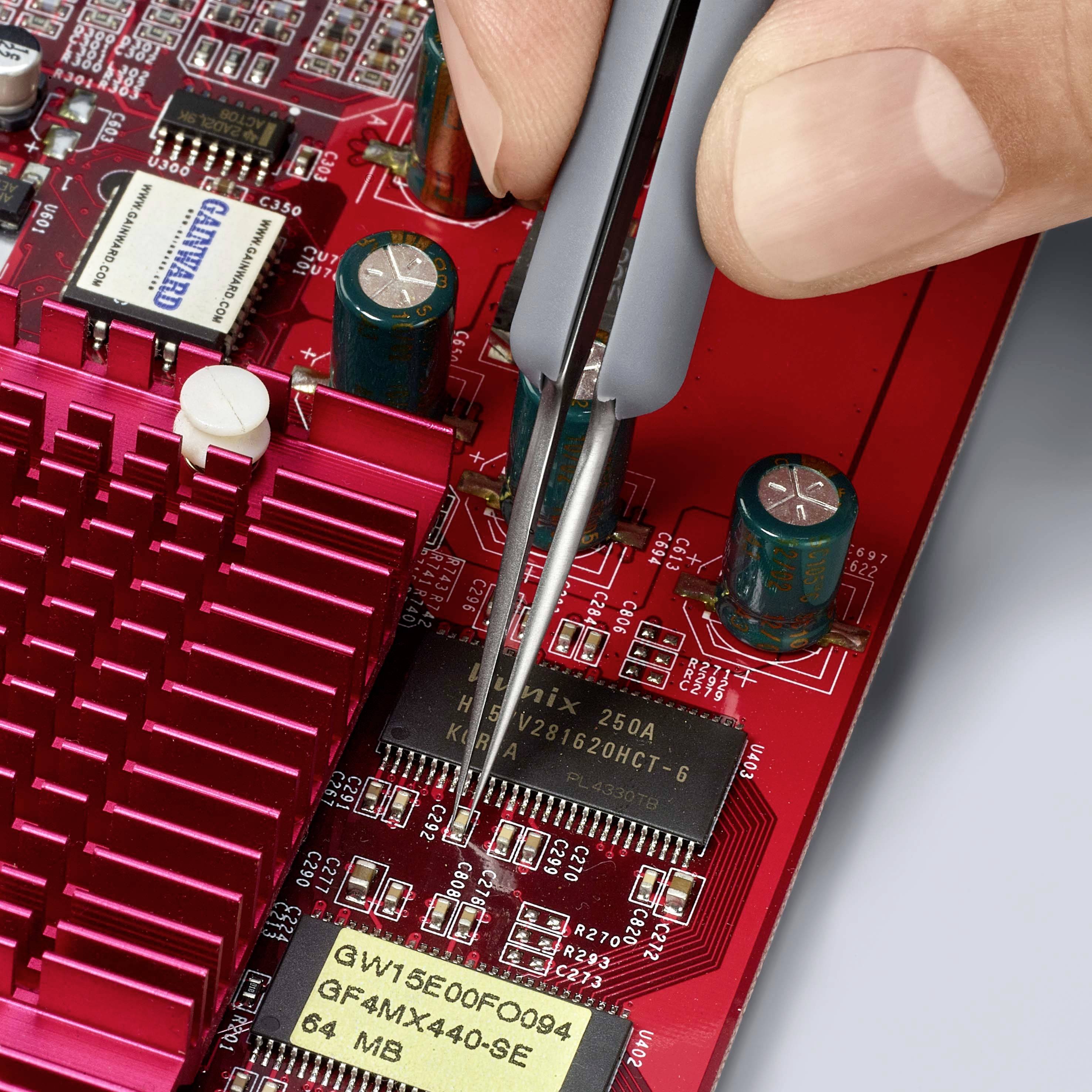 Osoba drží pinzetu, aby umístila čip na desku plošných spojů. Deska vykazuje několik elektronických součástek a označení.