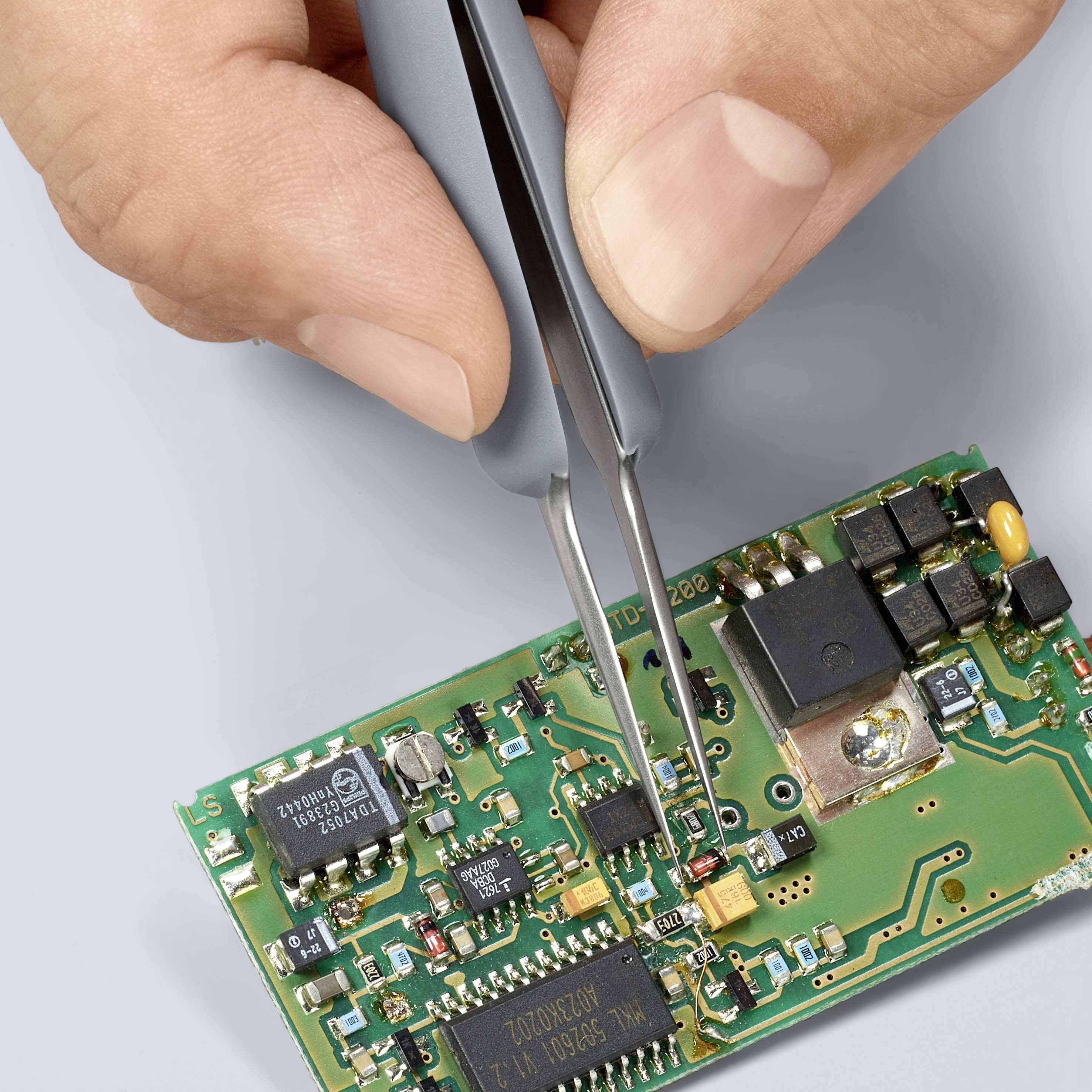 Jedna ruka drží pinzetu a umisťuje malou elektronickou součástku na zelenou desku plošných spojů s různými elektrickými obvody.