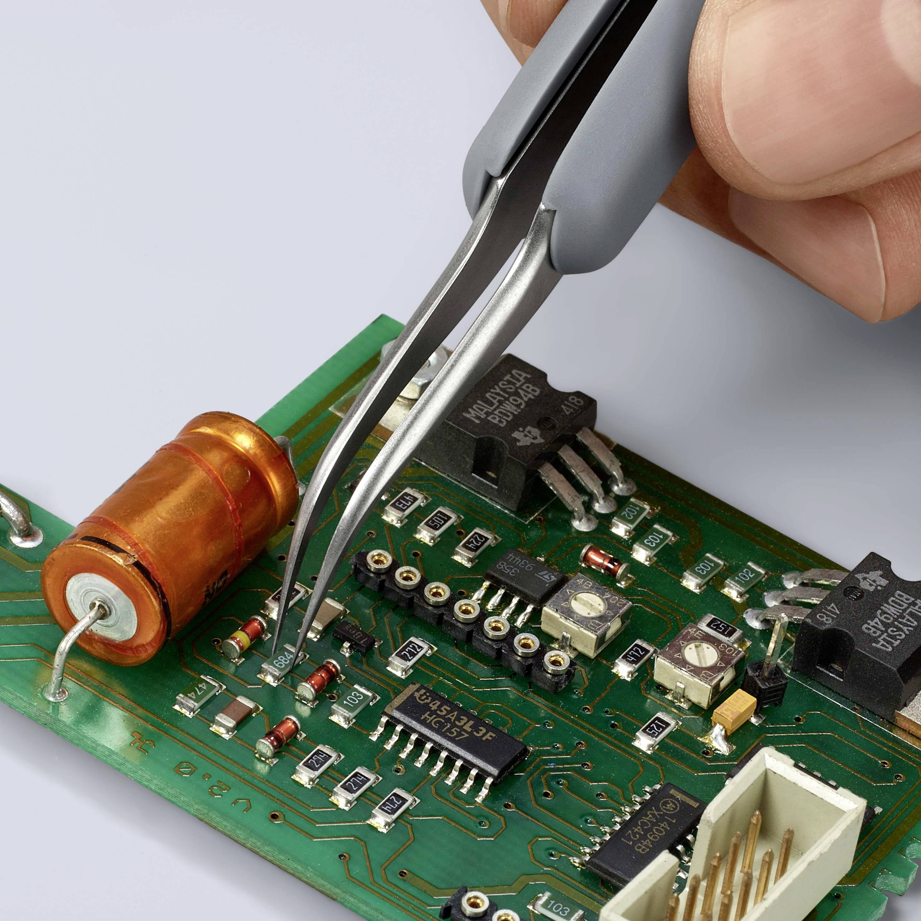 Jedna ruka drží pinzetu a umisťuje malou elektronickou součástku na zelenou desku plošného spoje osázenou čipy.