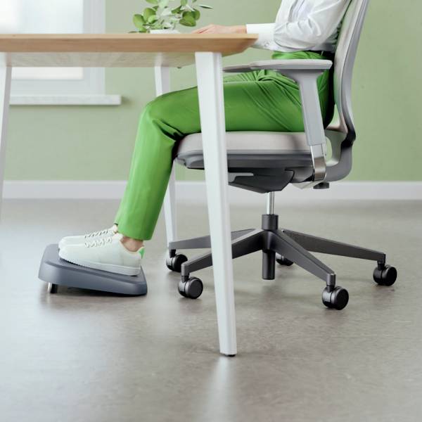 Osoba sedí na kancelářské židli u stolu a používá ergonomický trenažér nohou. V pozadí je zelená stěna.