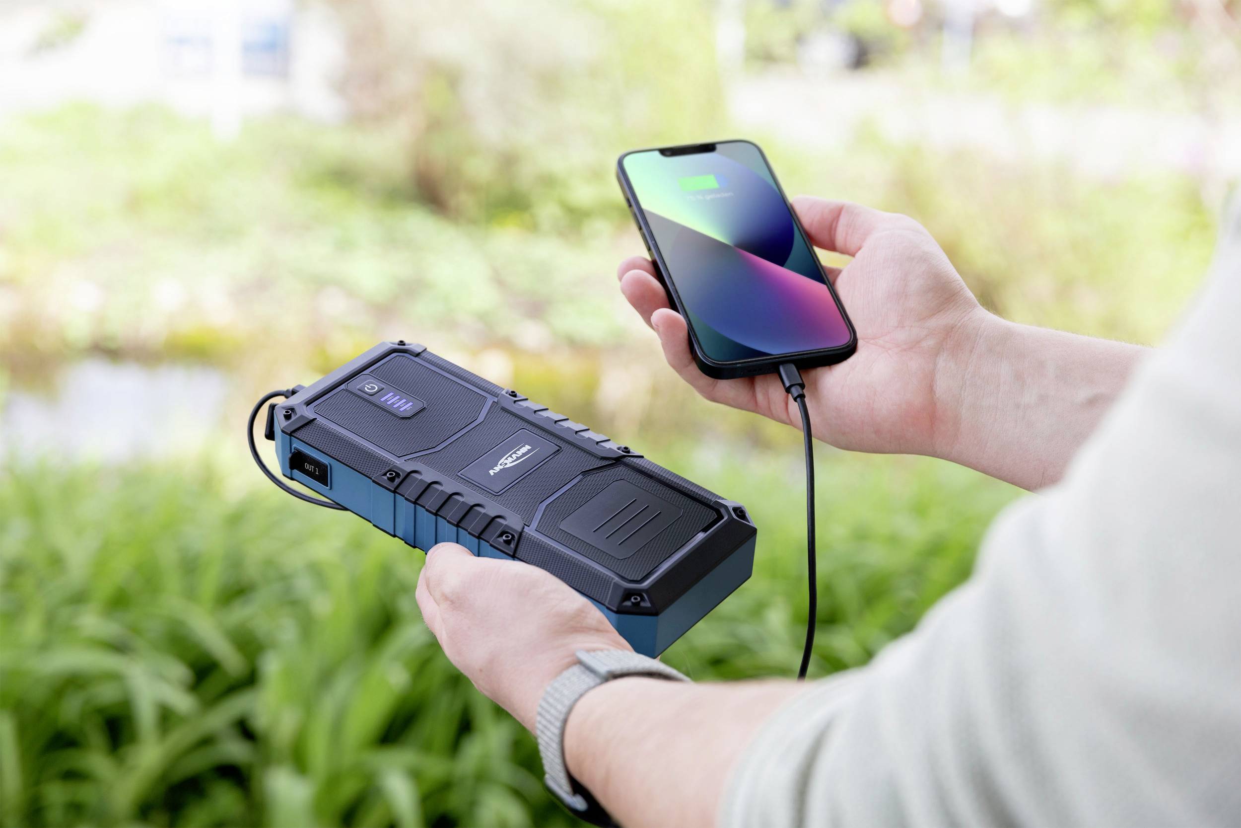 Osoba drží powerbabu a nabíjí smartphone venku. V pozadí je rozmazaná příroda.