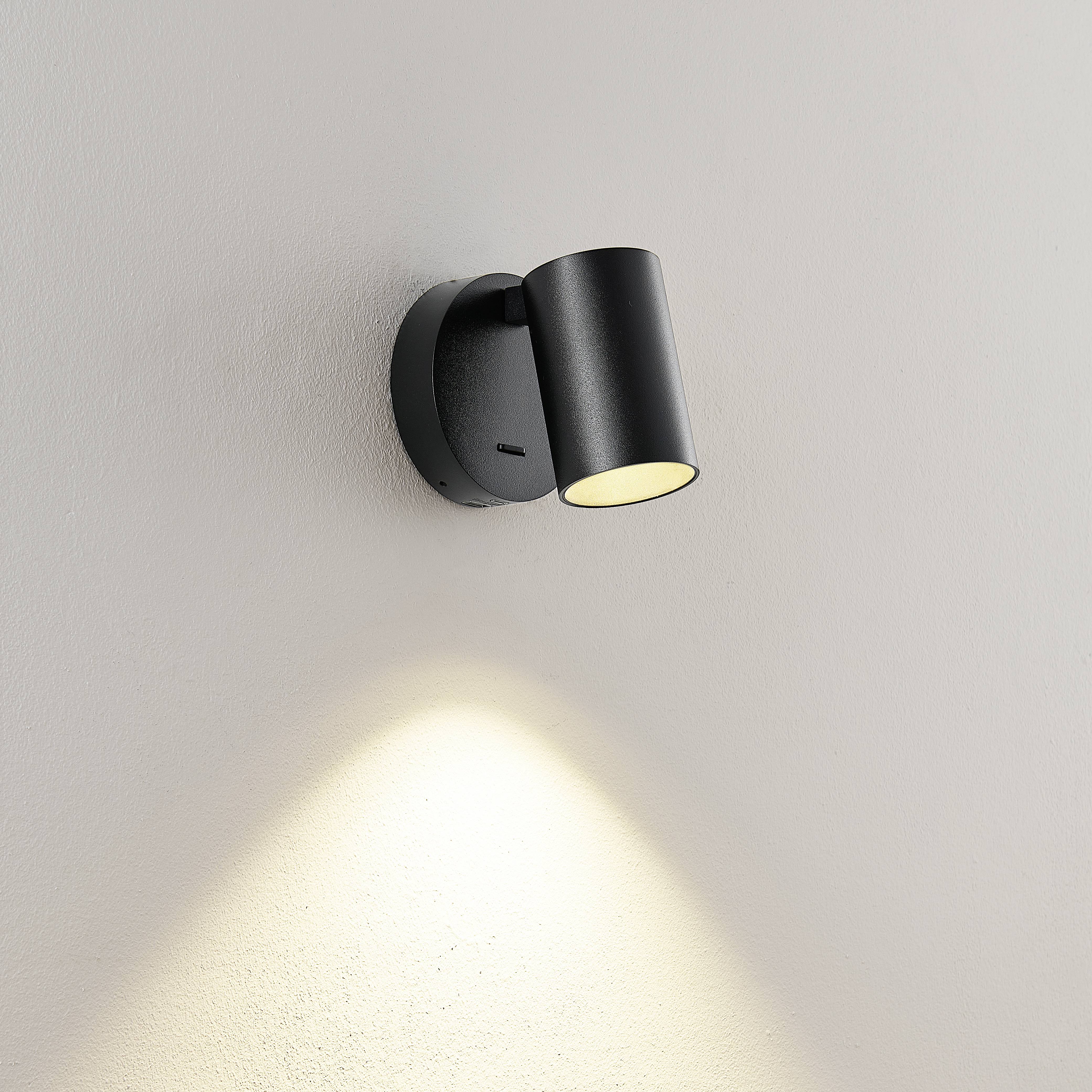 Černá nástěnná lampa s válcovým stínidlem, která směřuje teplé světlo dolů na bílou stěnu.
