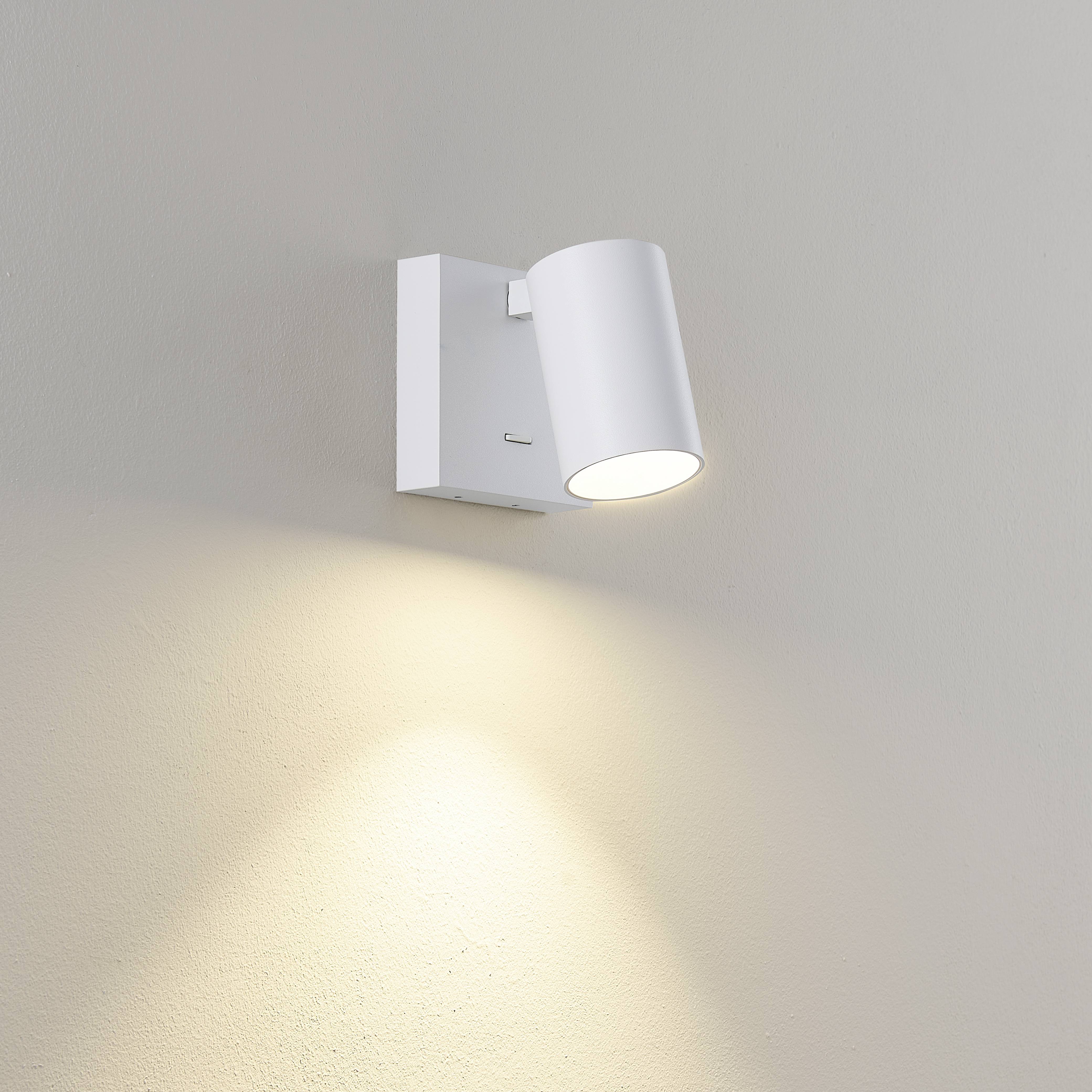 Bílá nástěnná lampa vyzařuje teplé světlo dolů a rovnoměrně osvětluje stěnu. Moderní, minimalistický design.
