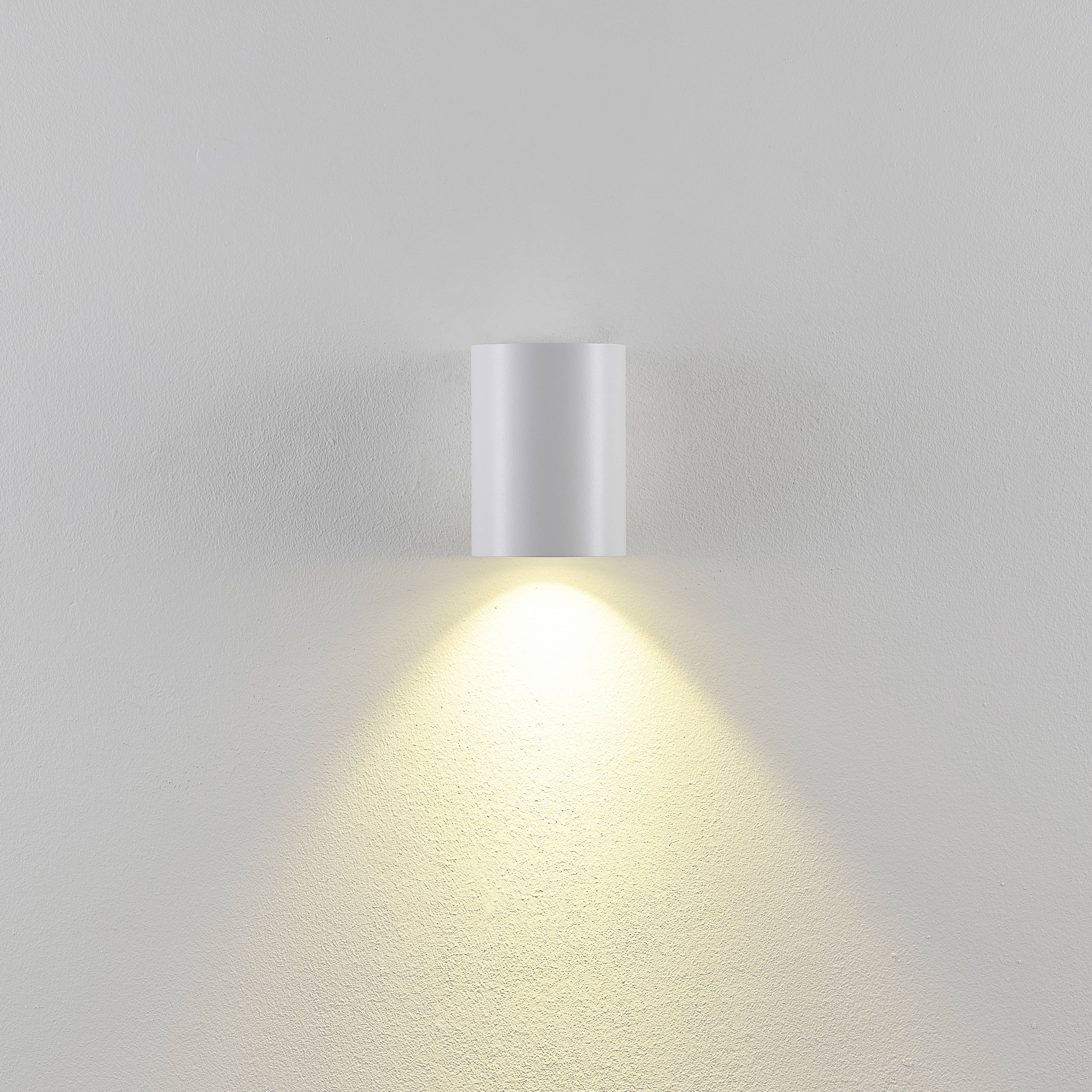 Bílá nástěnná lampa svítí dolů a vytváří teplý světelný kužel na šedé stěně. Minimalistický, moderní styl.