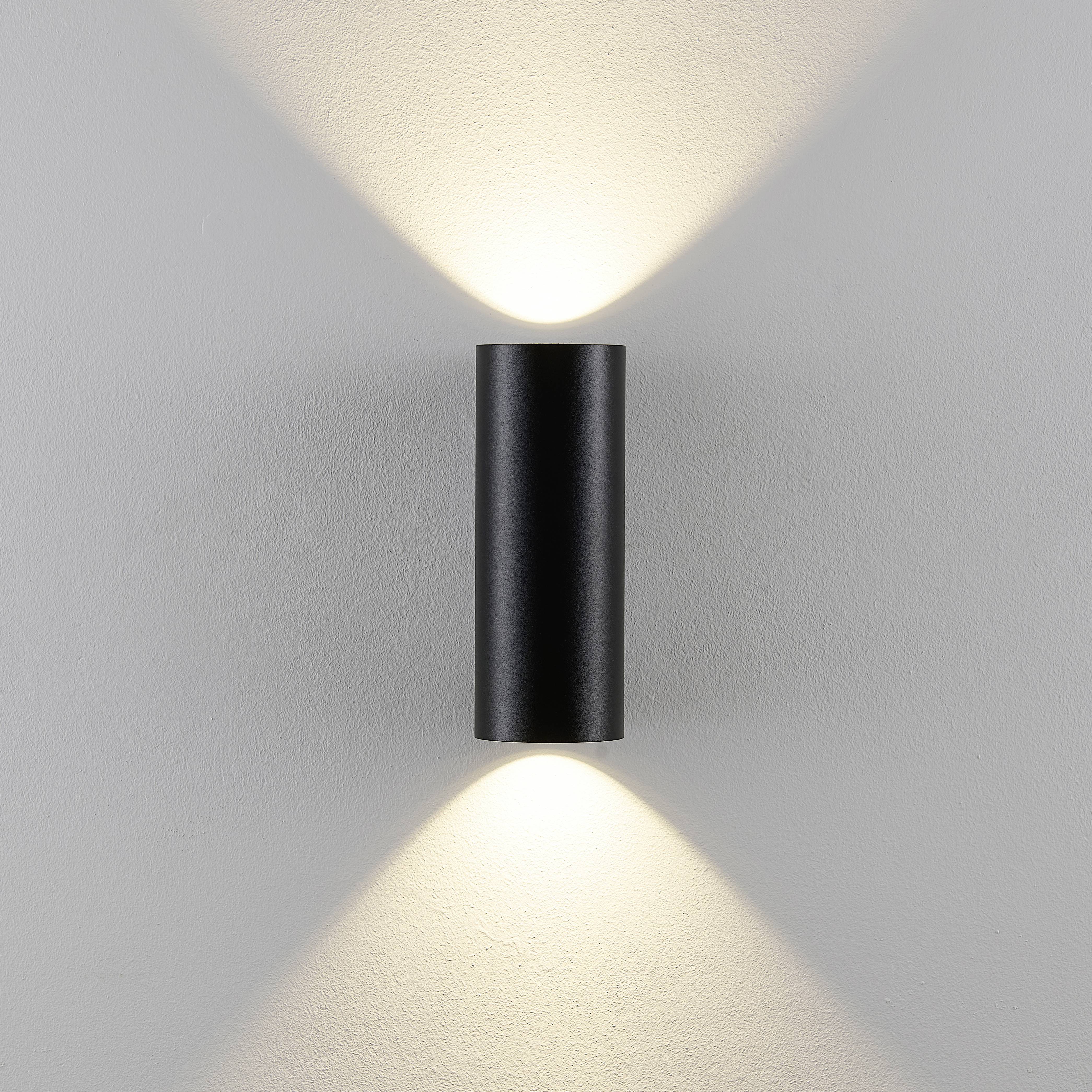 Nástěnná lampa válcovitého tvaru osvětluje bílou stěnu světlem směřujícím nahoru a dolů.