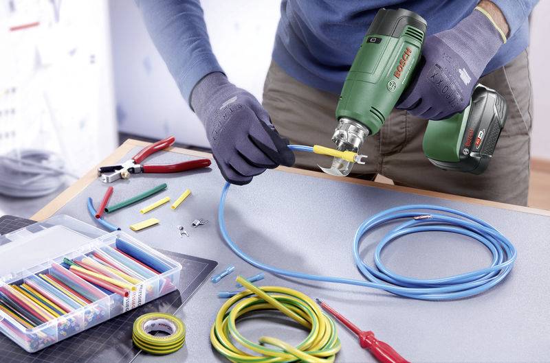 Elektrikář pracuje na kabelu s vrtačkou, kleštěmi a kabelovými konektory na stole, obklopen pracovními nástroji.