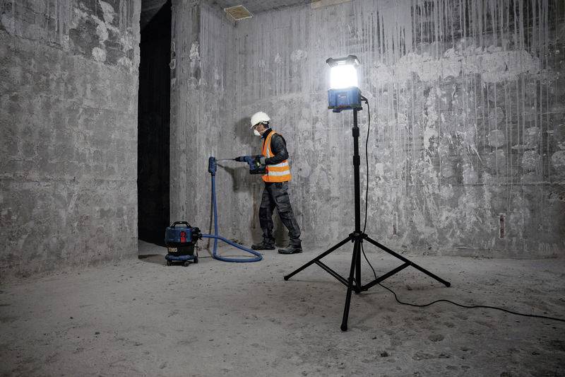 Dělník v bezpečnostním oblečení a helmě vrtá do betonové zdi, zatímco jasná lampa osvětluje prostor.