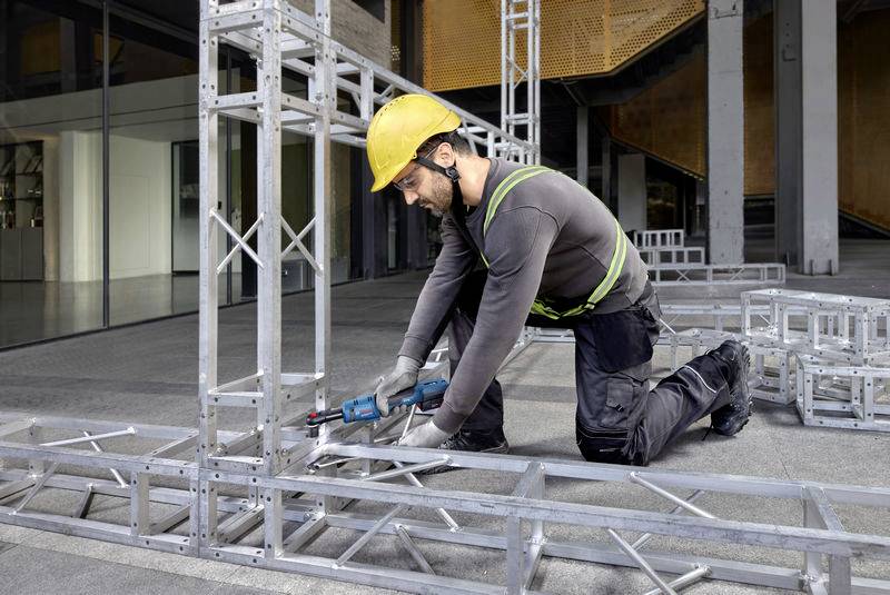Dělník s žlutou přilbou montuje velkou kovovou konstrukci pomocí elektického nářadí. Bezpečnostní vybavení je dobře viditelné.