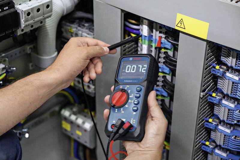 Jedna osoba měří napětí digitálním multimetrem v rozvaděči. Display ukazuje '0.72'.