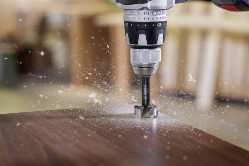 Vrtačka prochází dřevěnou deskou, třísky létají všemi směry. Pozadí je rozmazané.